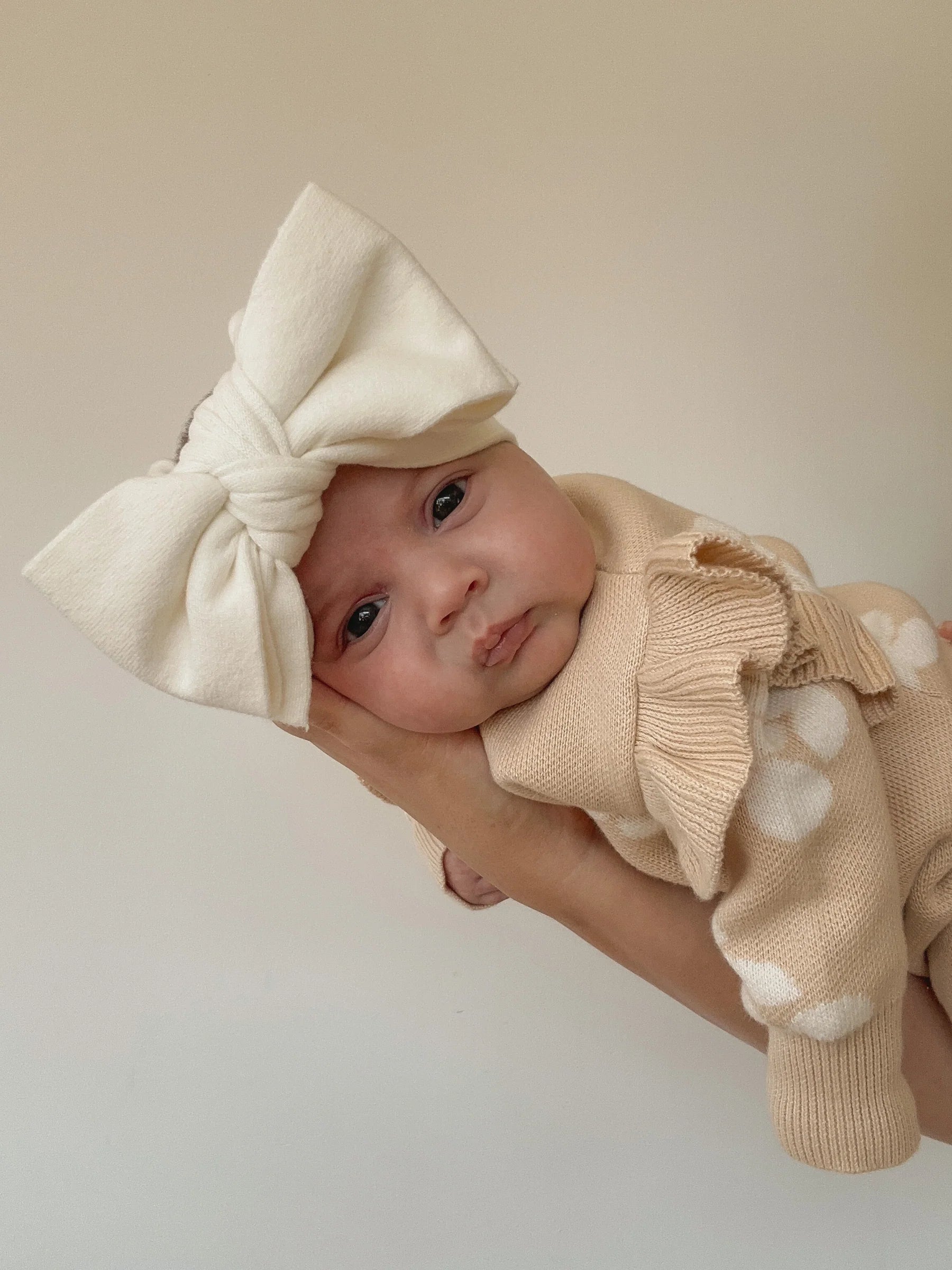A baby in a beige polka dot outfit wears the ZIGGY LOU Ziggy Lou - Headband Milk, gently held by an adult hand to highlight Baby Safety against a plain background.