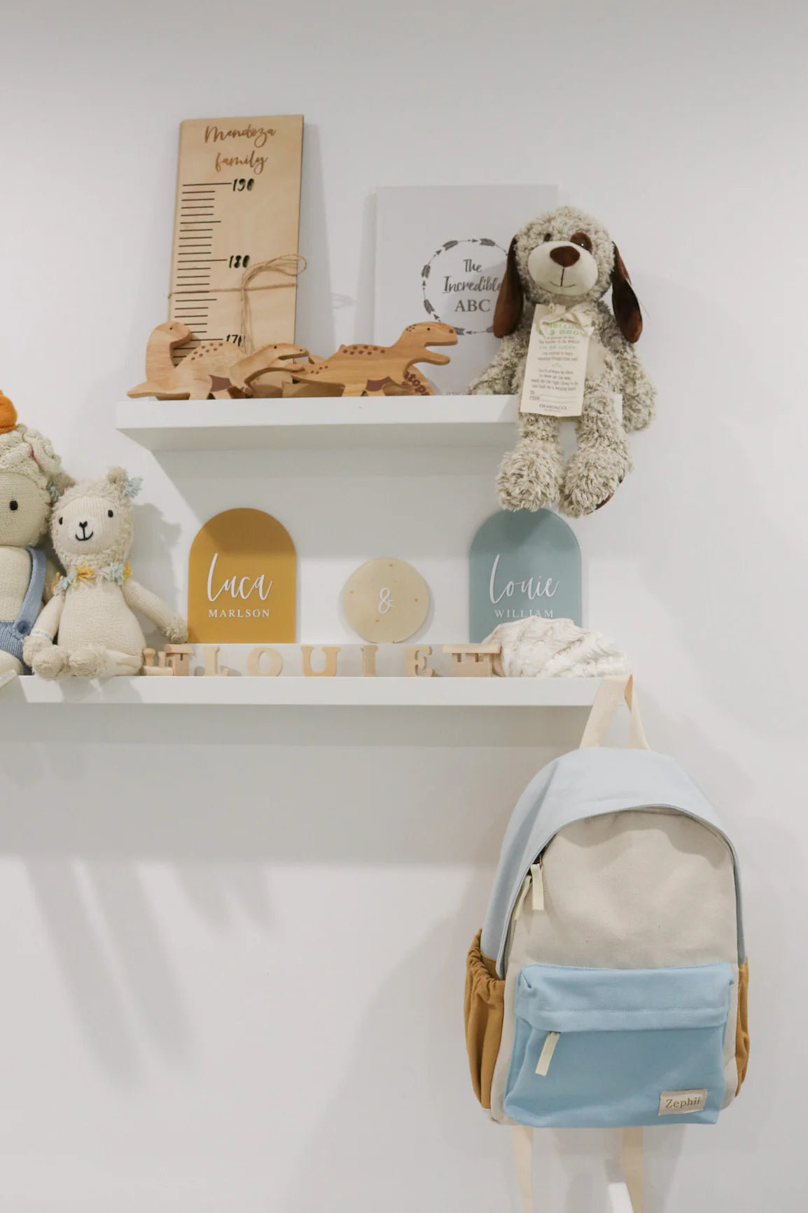 A children's room display showcases two white shelves brimming with stuffed animals, wooden toys, and a height chart. The Jagger Backpack by ZEPHII, featuring adjustable straps for personalization, is hung on the wall. The bottom shelf is adorned with name plaques and blocks for an added touch of charm.