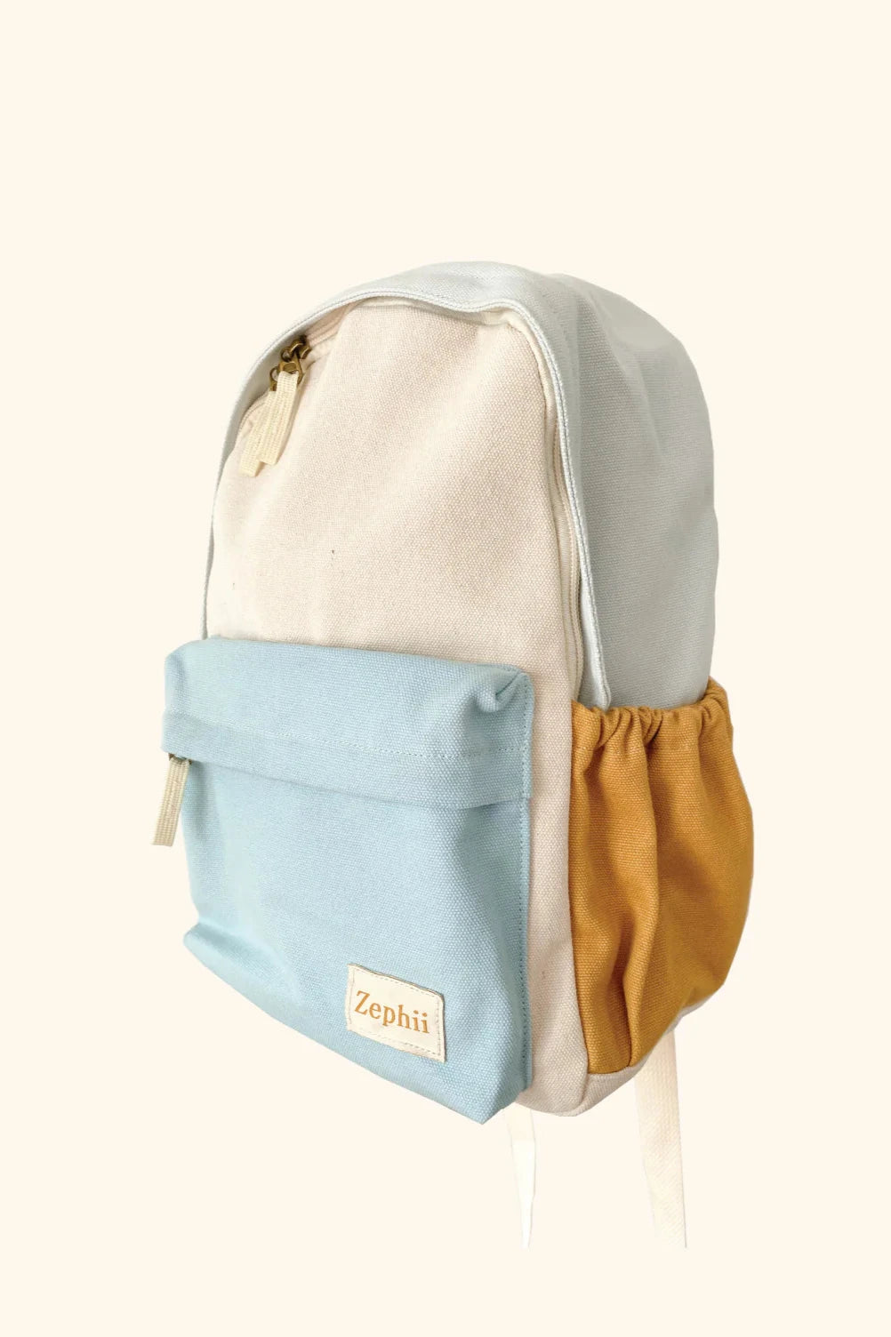 Introducing the Jagger Backpack by ZEPHII: This chic kindergarten bag comes in a sophisticated beige color with a light blue front pocket and a mustard side pocket. It includes adjustable straps for comfort and style, while a small beige tag displaying the brand name "ZEPHII" adorns the front pocket. Set against a simple background, this backpack perfectly embodies the JAGGER style's elegant essence.