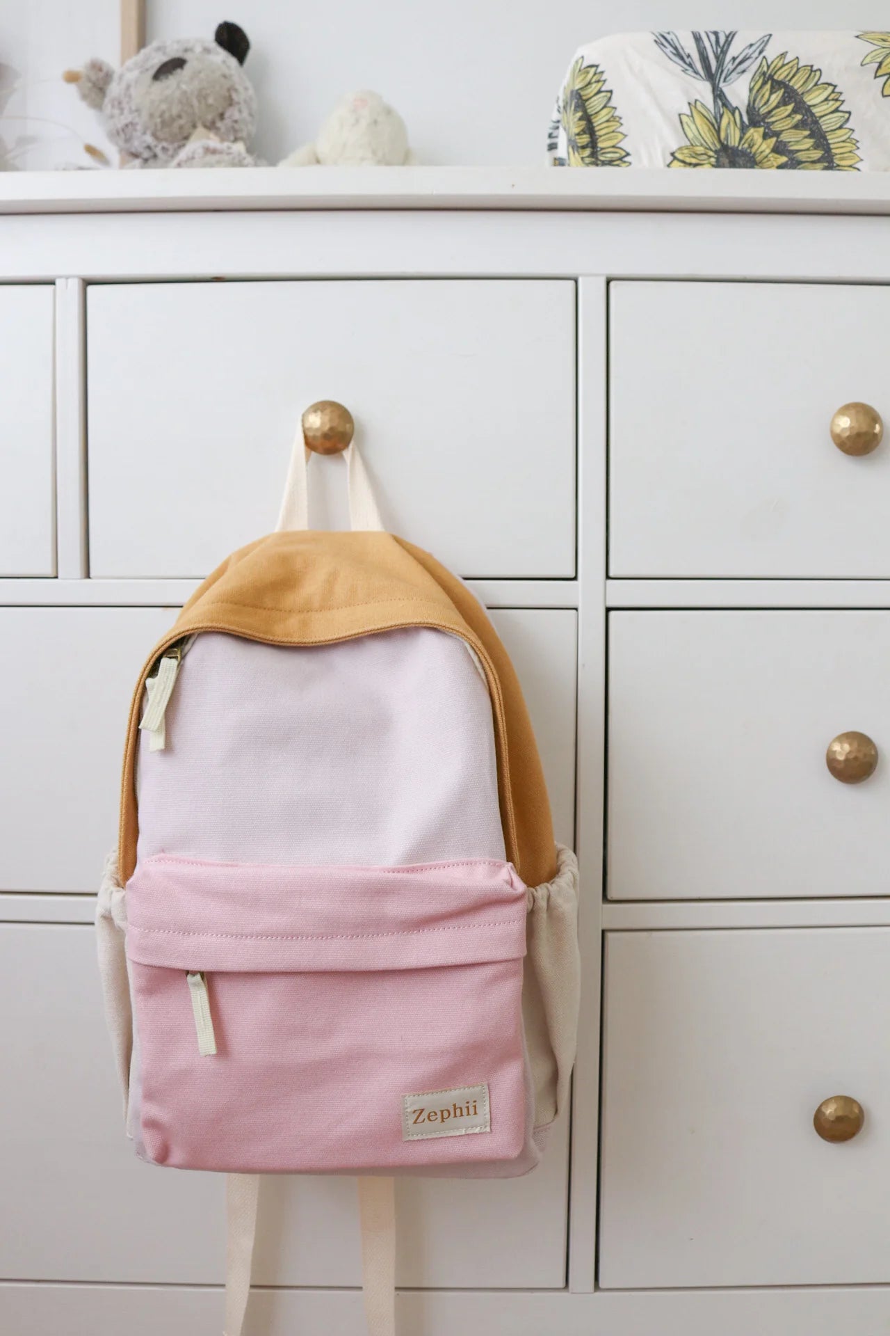 The ZEPHII Poppy Backpack, featuring tan, light pink, and beige panels, hangs on a white dresser with gold knobs while plush toys and a patterned item rest on top.