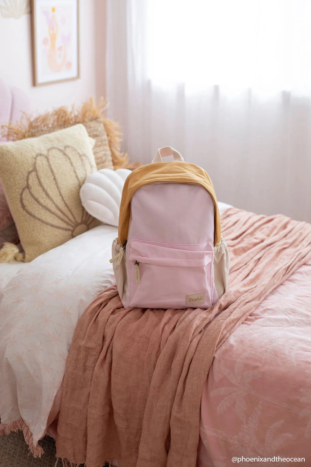 The ZEPHII Poppy Backpack in pink and beige is displayed on a neatly made bed with pink and white bedding, a shell-shaped pillow, and soft light filtering through sheer curtains.