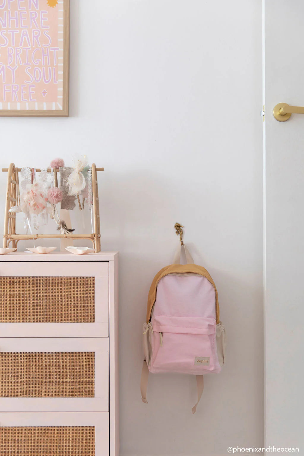 A ZEPHII Poppy Backpack in pink hangs from a wall hook next to a dresser with woven drawers. Above, a hair accessory rack and a framed inspirational quote add charm to the white wall.