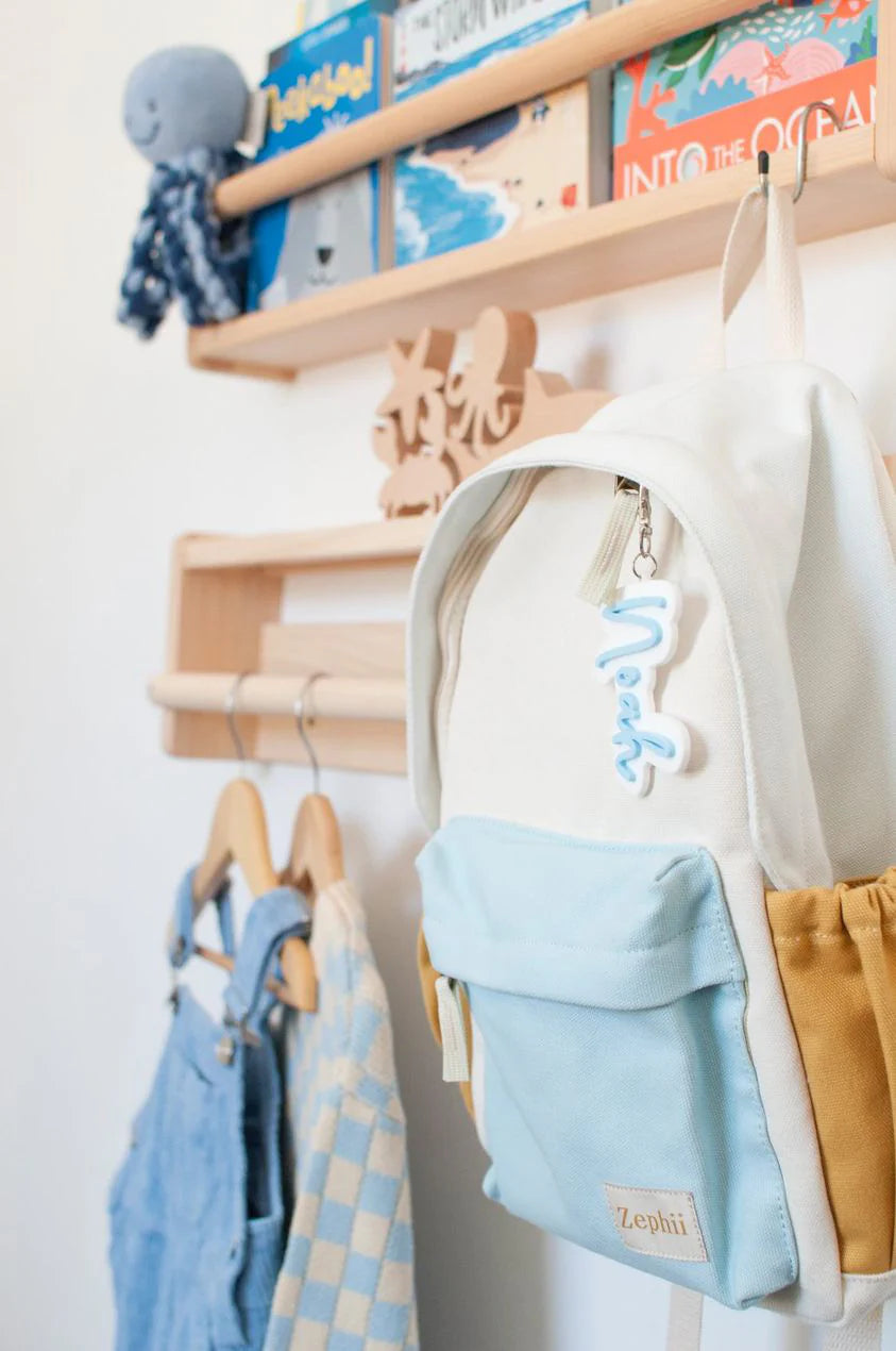A neatly organized wooden wall shelf displays children's books and a small blue octopus toy. Below, a Jagger Backpack by ZEPHII with adjustable straps and a "Neal" keychain hangs alongside blue and yellow children's clothing on hangers.