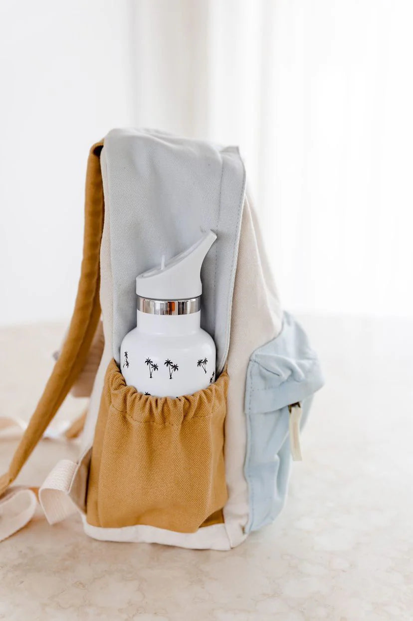A light blue and beige Jagger backpack from ZEPHII rests on the table, showcasing a side pocket holding a white water bottle decorated with small black palm tree designs and finished with a metallic lid. This bag comes equipped with adjustable straps and a handy front zipper pocket.