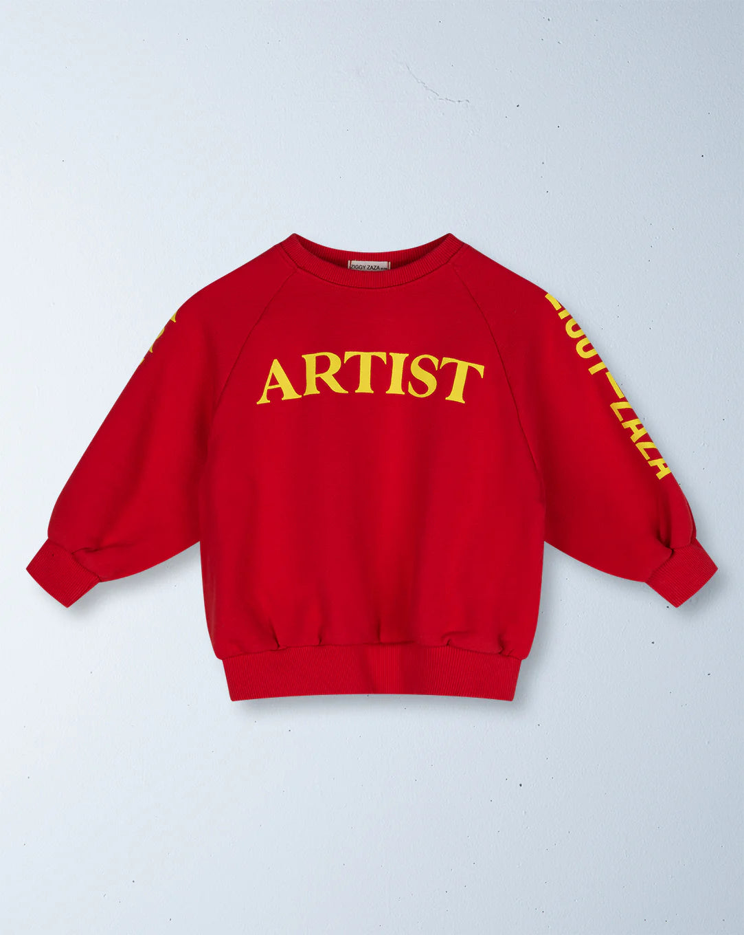 The ZIGGY ZAZA Ziggy Zaza - Artist Pullover Racer Red features a relaxed fit, bold yellow "ARTIST" text on the chest, partial yellow sleeve graphics, and is made from 100% cotton fleece, displayed against a light gray wall.