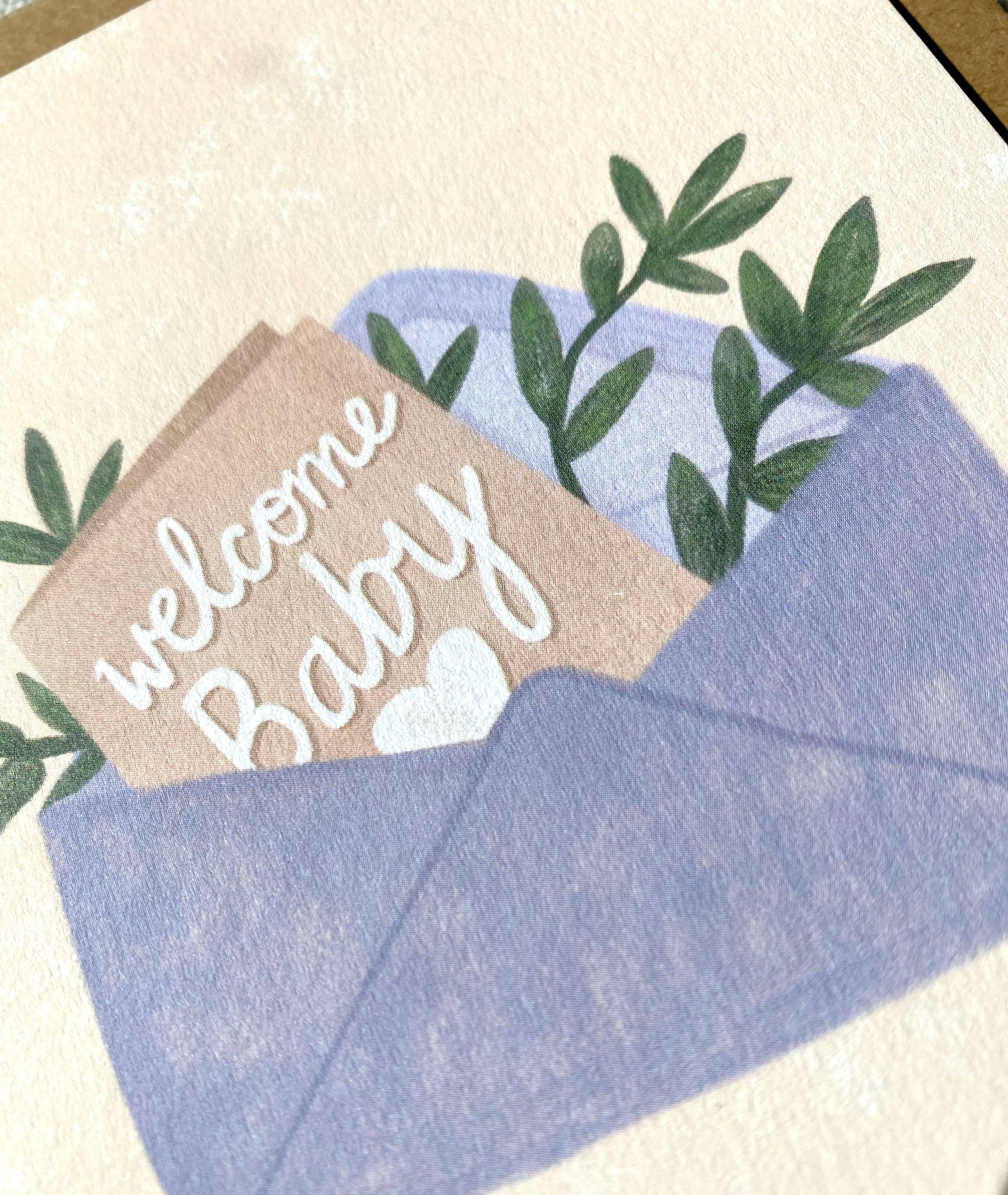 An open blue envelope reveals a "Welcome Baby" gender-neutral card by Lauren Sissons Studio, crafted from recycled paper. It features a white heart and green leafy branches peeking out.