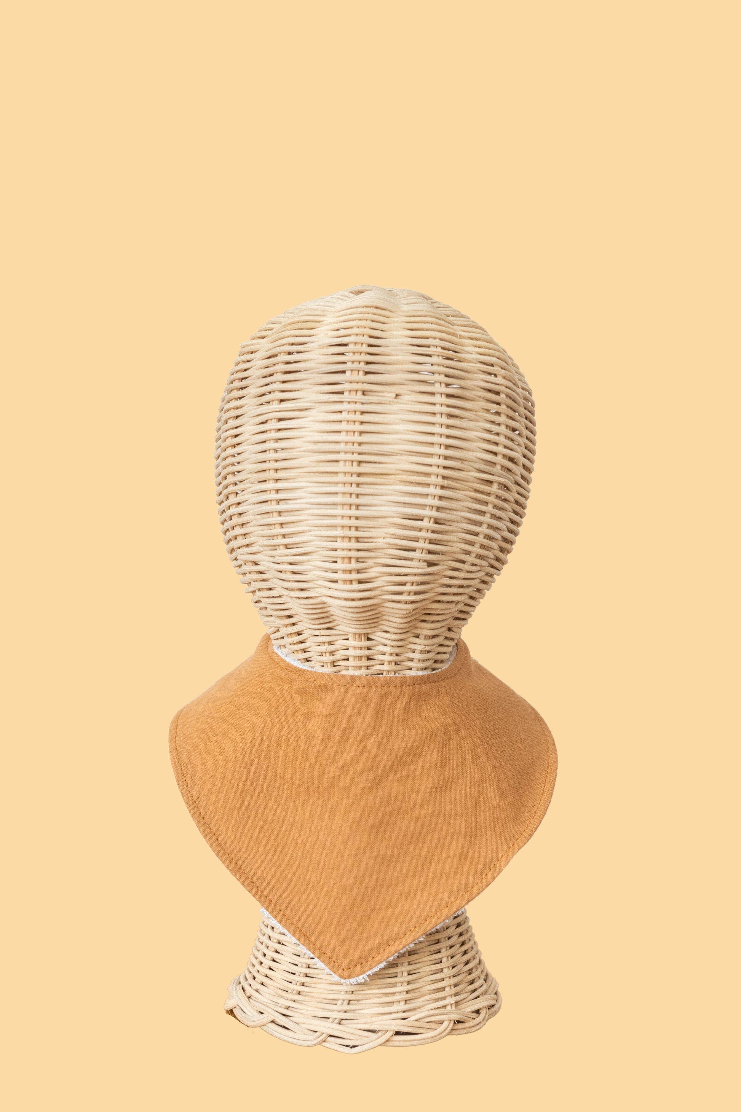 A wicker mannequin displays the Kiin Baby Dribble Bib, featuring a brown bandana made from an absorbent bamboo/cotton blend, set against a solid beige background.