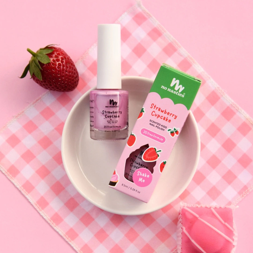 A bottle and box of NO NASTIES No Nasties - Scented Scratch Off Kids Nail Polish Strawberry Cupcake - Pastel Pink rest in a white bowl on pink gingham, beside a strawberry and heart-shaped pastry—ideal for fun kids' nail polish play.