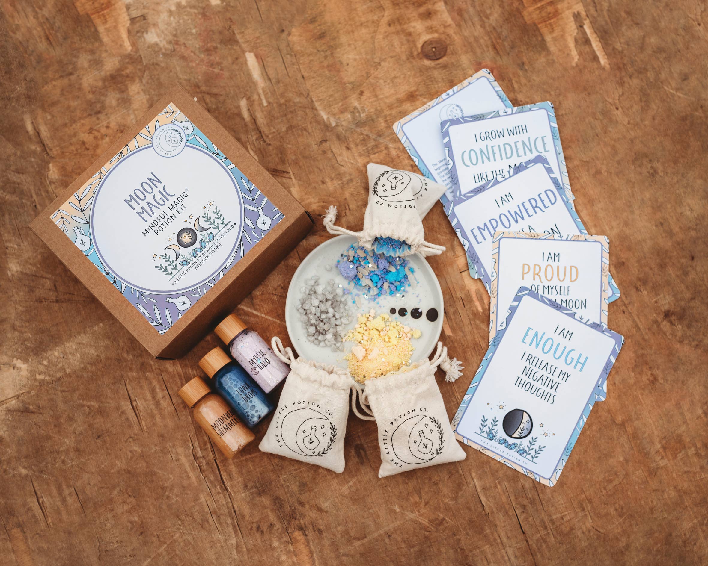 A wooden surface displays the "Moon Magic MINDFUL Magic Potion Kit" by THE LITTLE POTION CO, showcasing a mindfulness activity with a box, small bags of crystals, three bottles of colored powder, blue and yellow crystals on a white plate, and affirmation cards featuring positive messages like "I AM PROUD OF MYSELF.