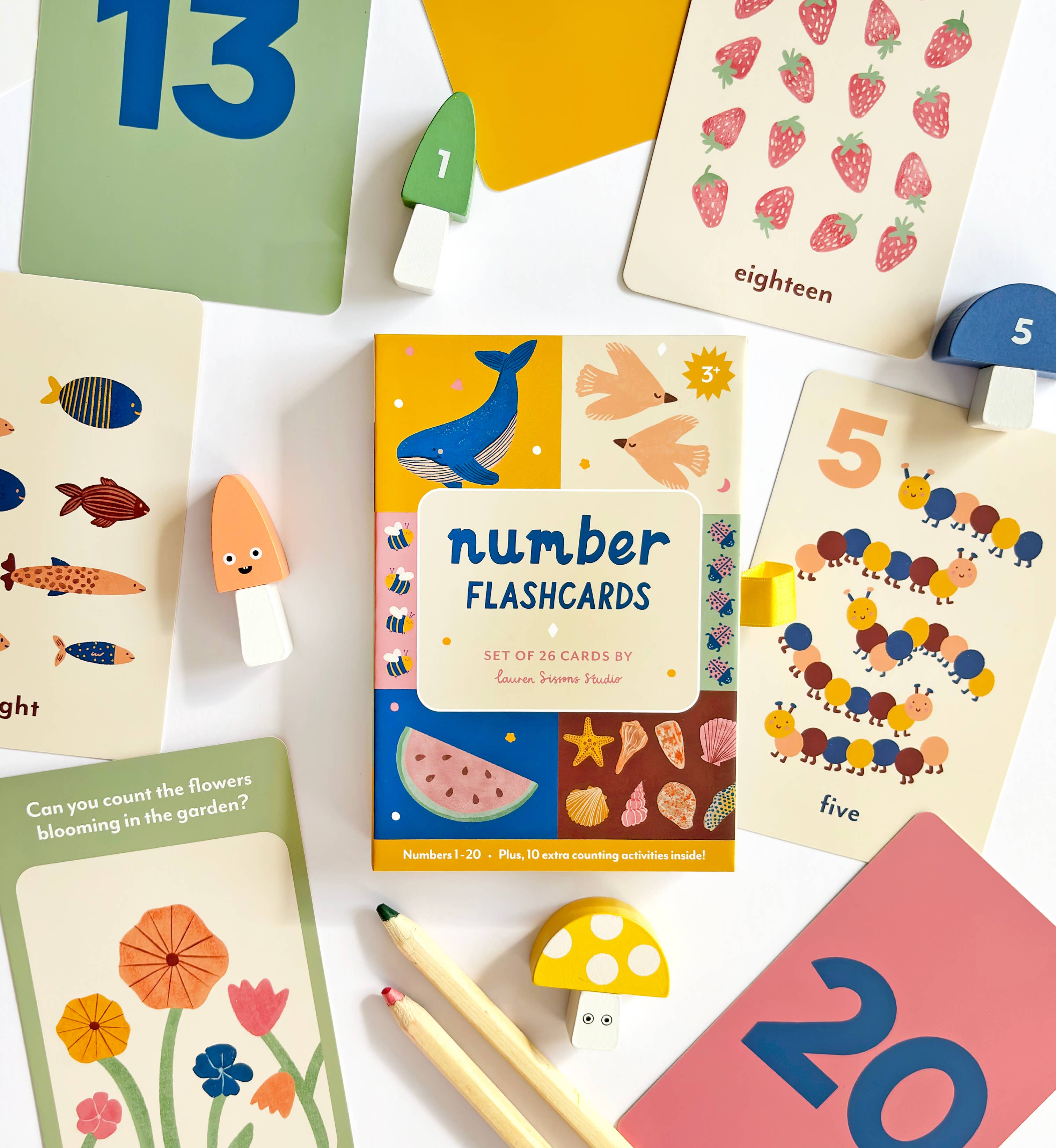 Lauren Sissons Studio's Illustrated Number Flashcards are spread out on a white surface, featuring colorful animal, plant, and object illustrations to teach counting. Wooden mushrooms and pencils are also in view.