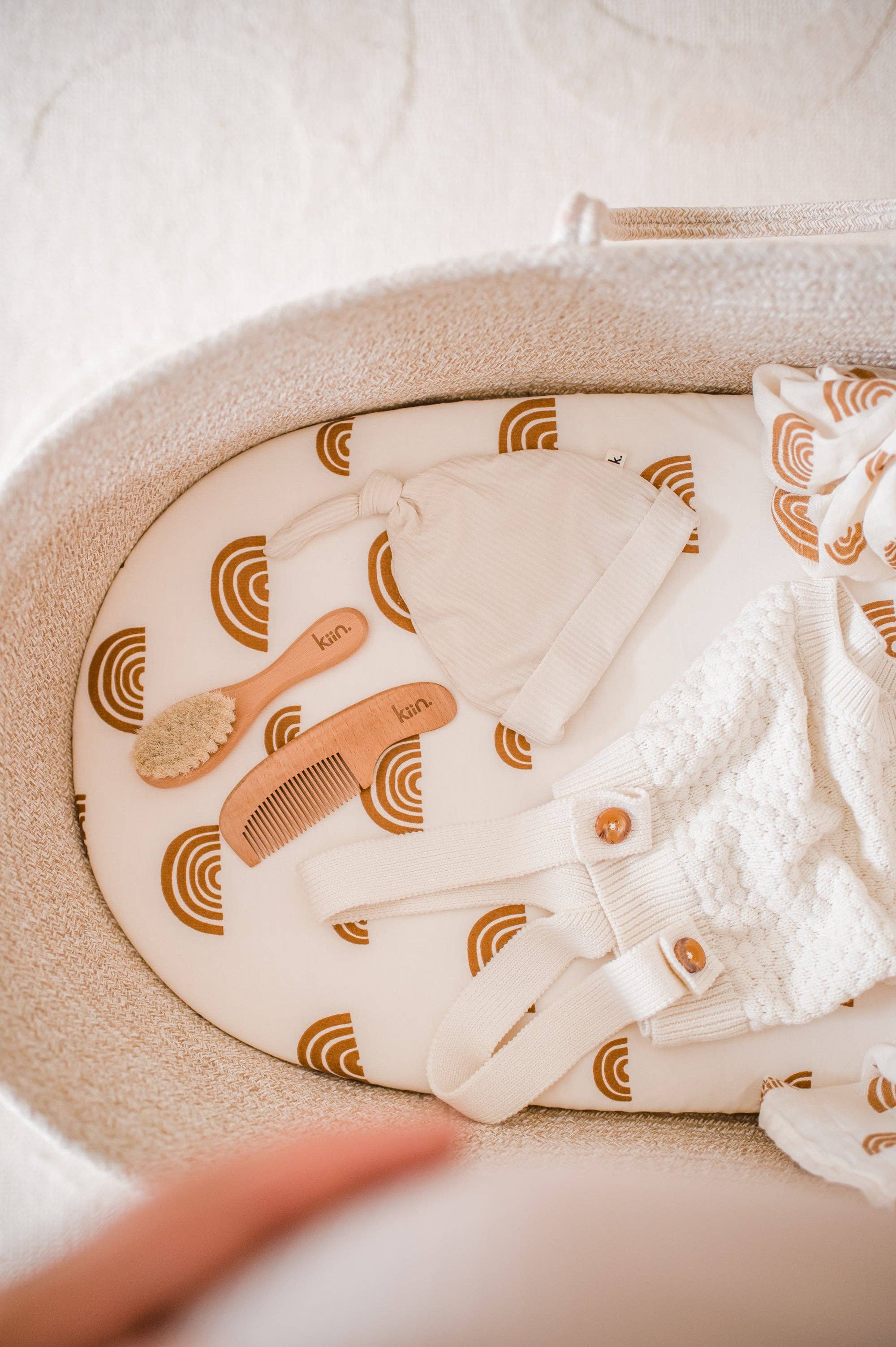 A baby bassinet with a patterned mattress featuring brown arches holds a cozy collection: a white baby beanie, KIIN BABY's Wooden Baby Brush and Comb Set, and white knitted baby clothes with wooden buttons. Each item is thoughtfully crafted to welcome your little one in style.