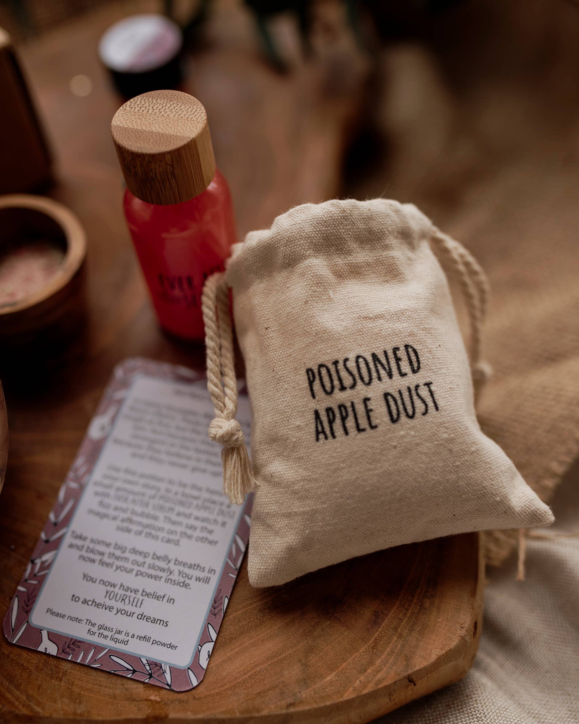 A small "Poisoned Apple Dust" drawstring pouch from The Little Potion Co's MINI Once Upon A Potion Kit sits on wood, with a red bottle and card nearby against an earthy-toned backdrop, inviting imaginative play.