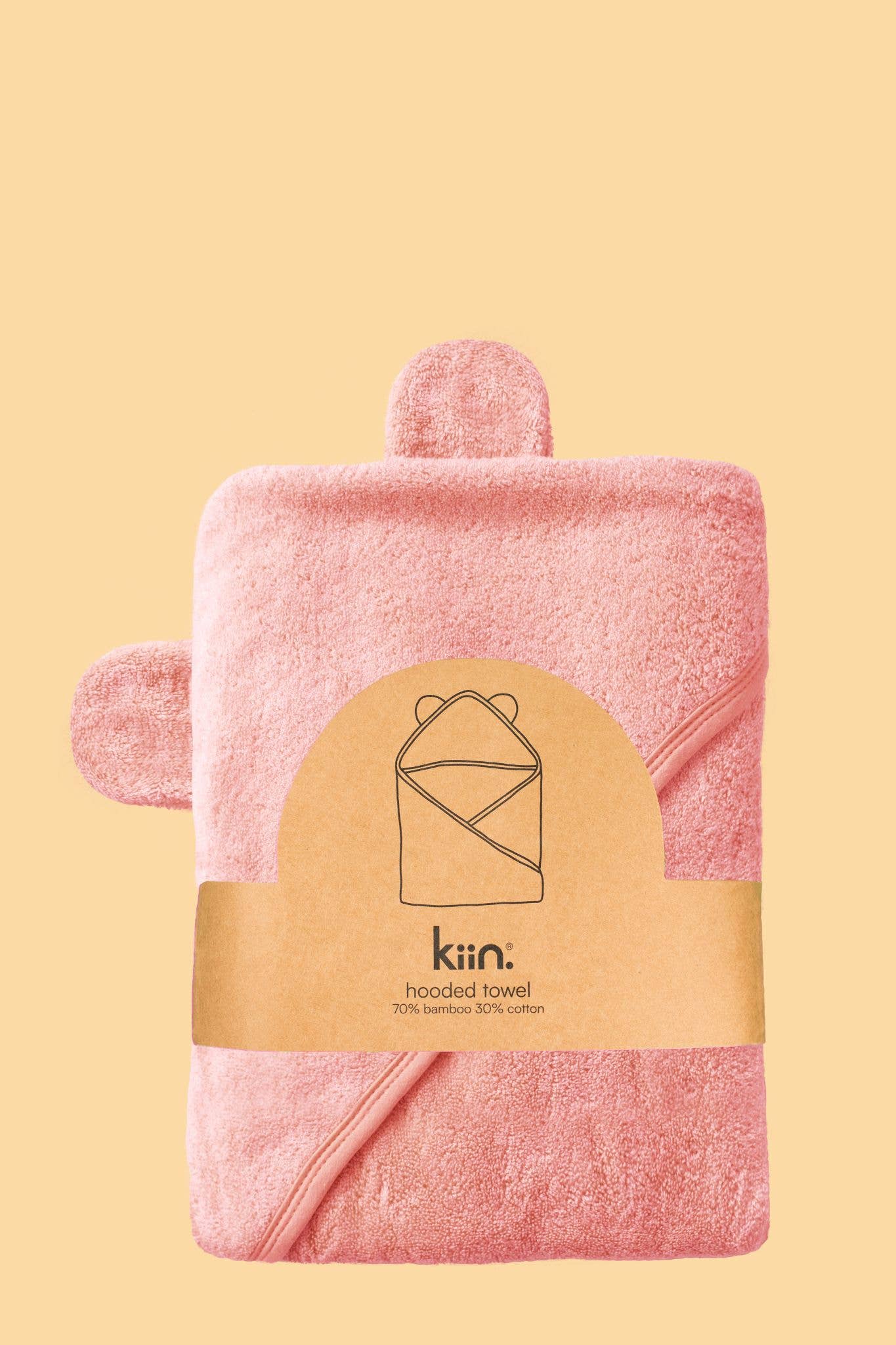 A soft pink hooded towel by KIIN BABY is neatly folded on a plain yellow background. The minimal packaging shows a line drawing and text stating it's crafted from 70% hypo-allergenic bamboo and 30% cotton for gentle comfort.