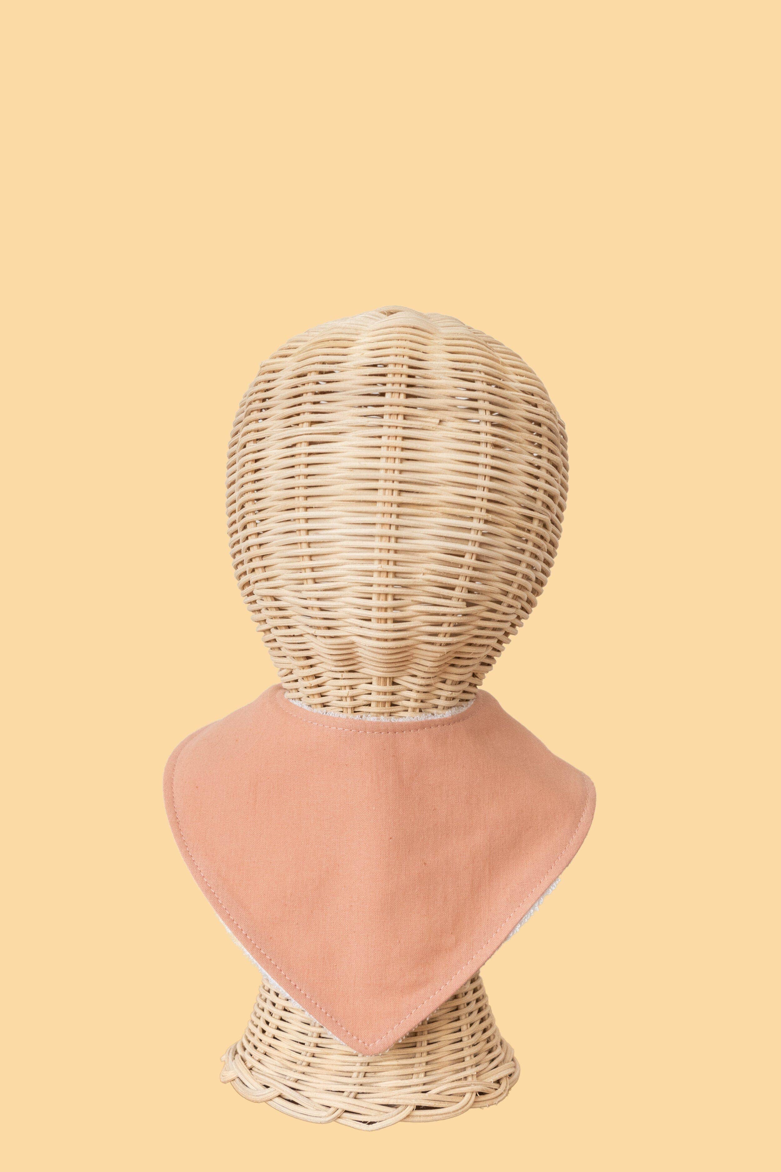 Displayed on a woven wicker mannequin head, the KIIN BABY Dribble Bib Dusty Rose is soft and peach-colored, made from absorbent bamboo/cotton towelling. It rests against a light beige background with a simple, pattern-free design.