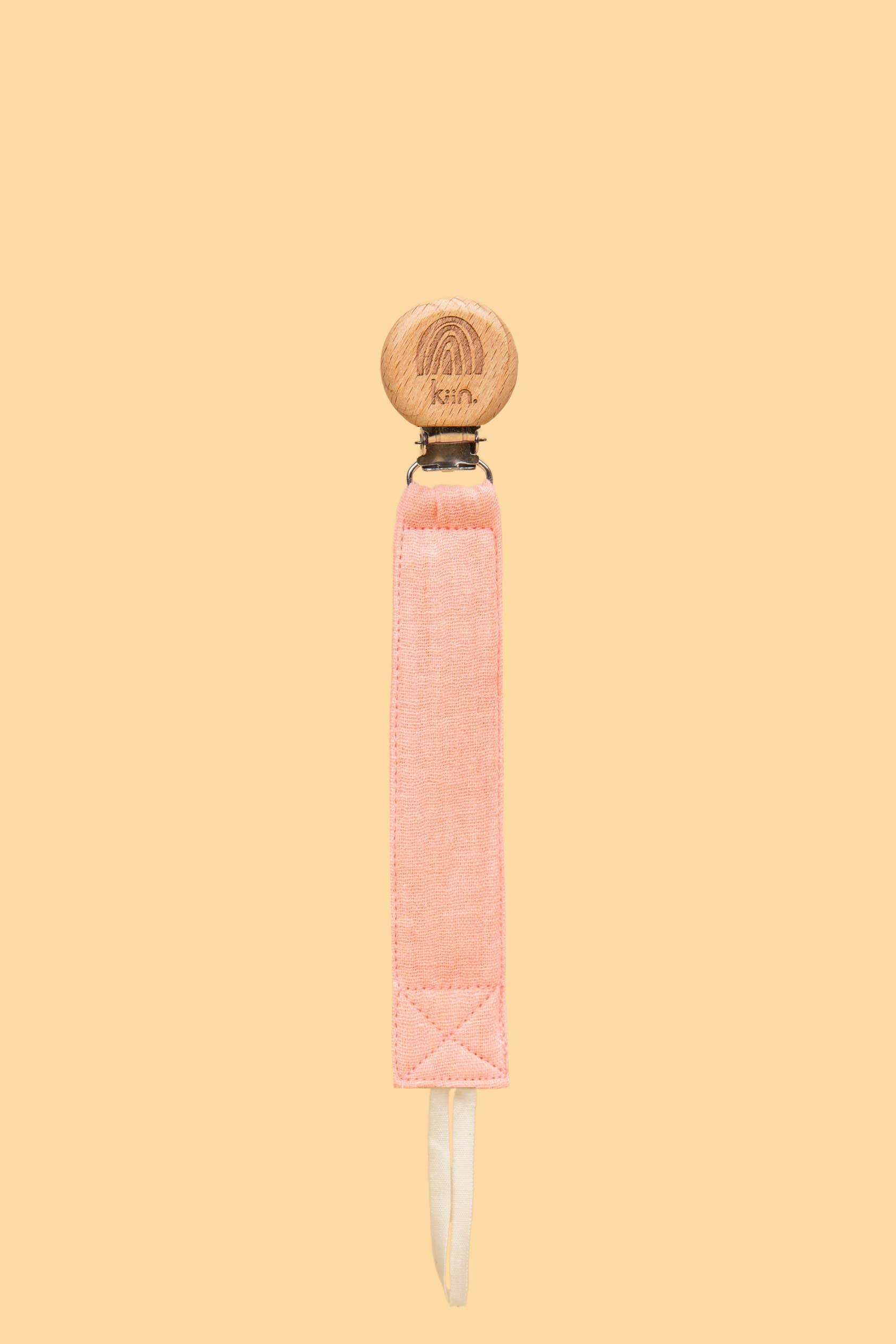 The Cotton Dummy Clip by Kiin Baby showcases a wooden pacifier clip with a carved rainbow and "krín" on top. It includes a pink cotton strap with a loop against beige, making it an ideal baby shower gift.