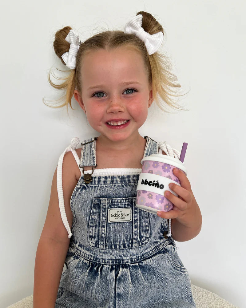 A young child with two white hair buns smiles while holding a BBCINO BBcino ~ Reusable BambinoCino Cup 240ml Blossom, featuring floral designs and a straw. They wear denim overalls over a white shirt, sitting against a plain background.