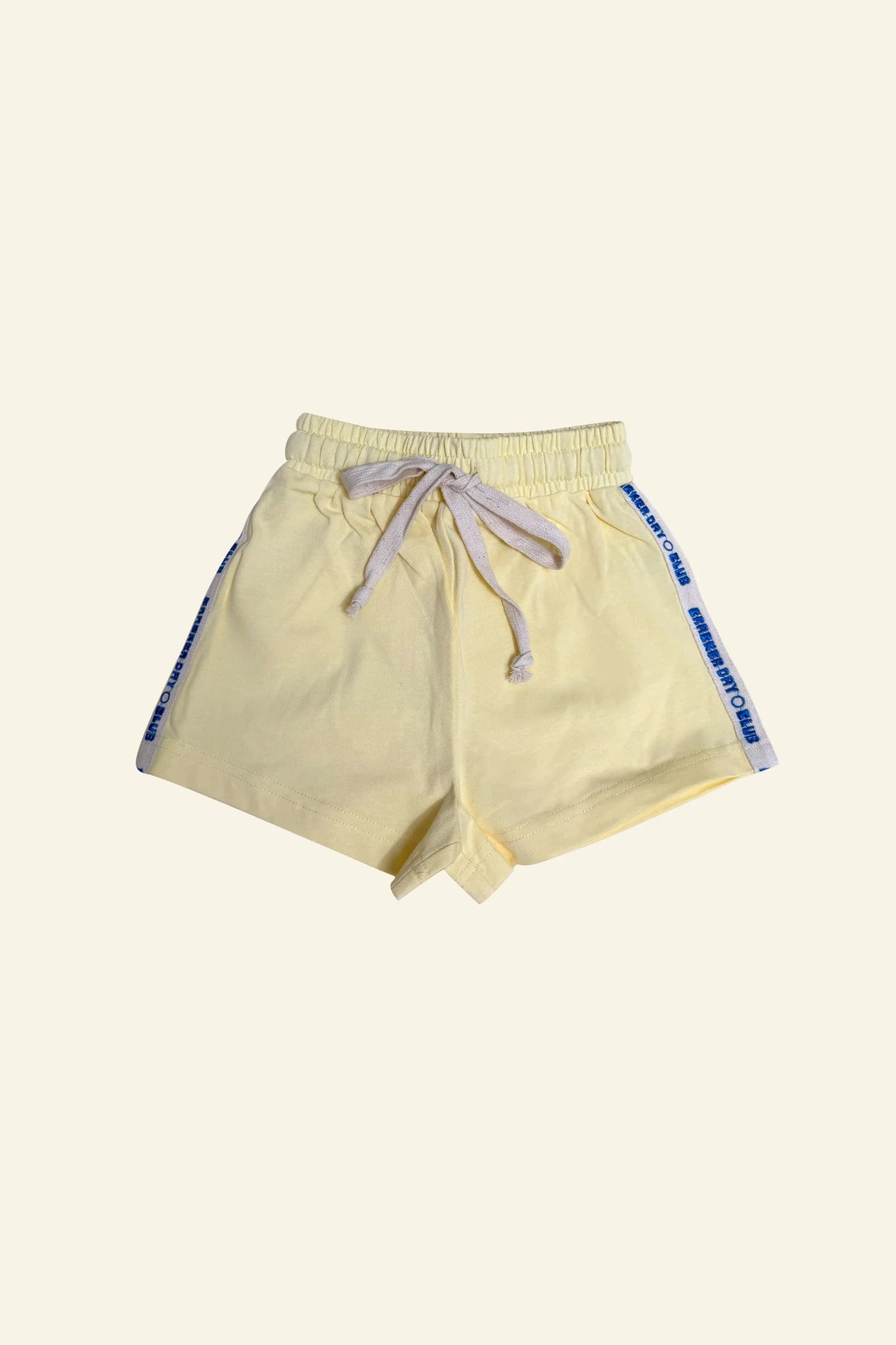 CRACKER DAY CLUB’s Kids Racer Club Shorts Sunshine feature a light yellow color, elastic waistband, white drawstring, and blue retro stripe accents with text. The cotton shorts are shown laid flat on a plain light background.