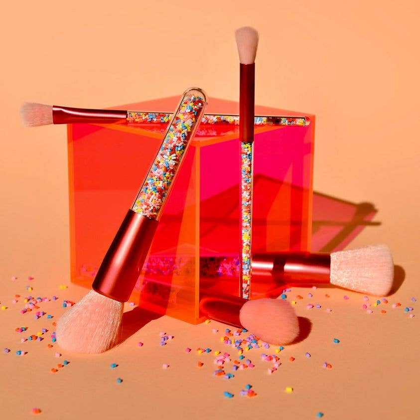 The Twinkle Sprinkle Brush Set by no nasties kids showcases vegan and cruelty-free makeup brushes boasting colorful, sparkly handles. These brushes are displayed around a translucent, red-orange acrylic cube. The hypoallergenic set features soft, fluffy pinkish bristles with multicolored sprinkles adorning the surface around both the brushes and the cube.