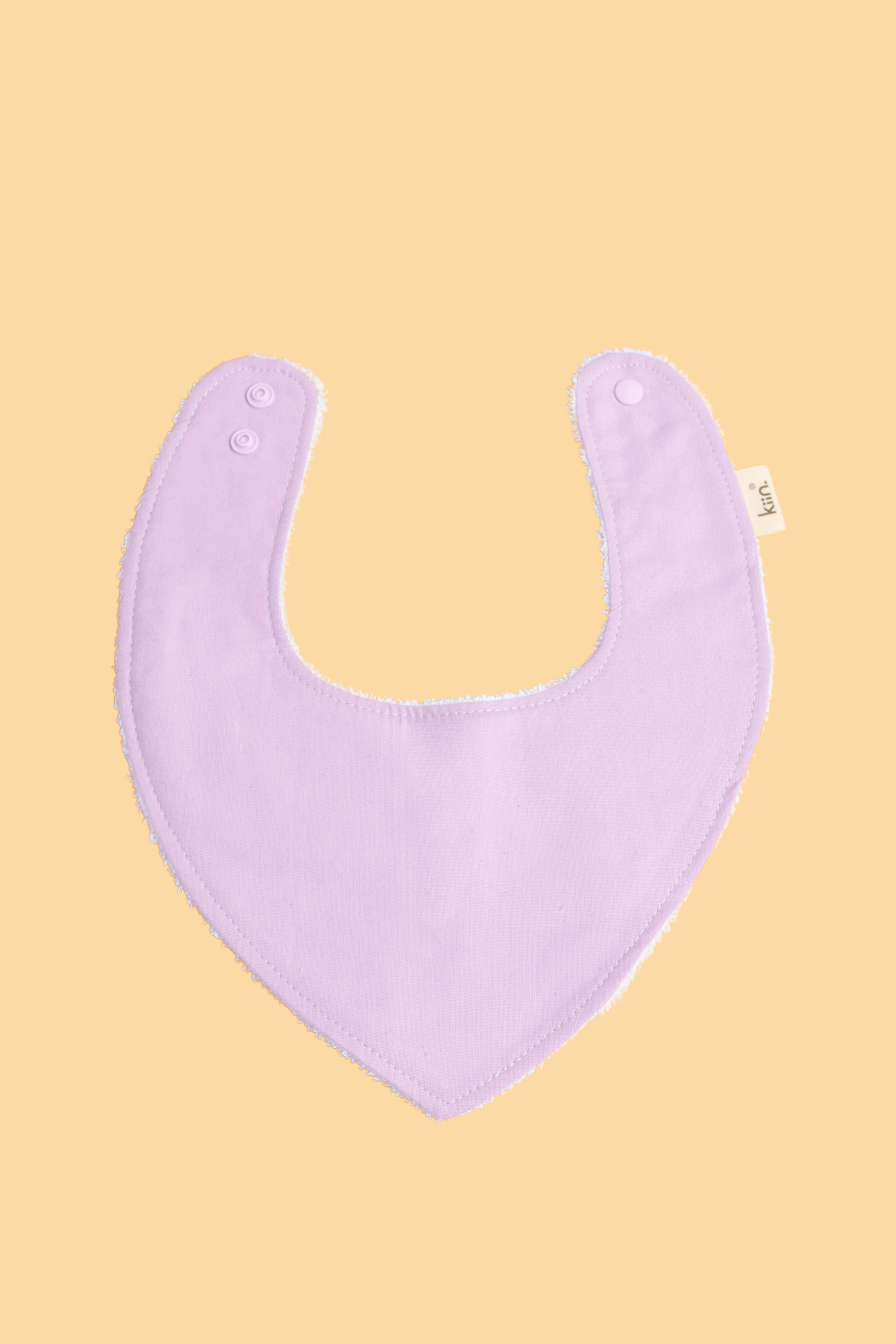 The Dribble Bib by Kiin Baby is a soft, lavender-colored bib with a U-shape crafted from a bamboo/cotton blend for added absorbency. It includes two snap buttons for adjustable fitting and a small fabric tag on the side, set against a warm beige background.