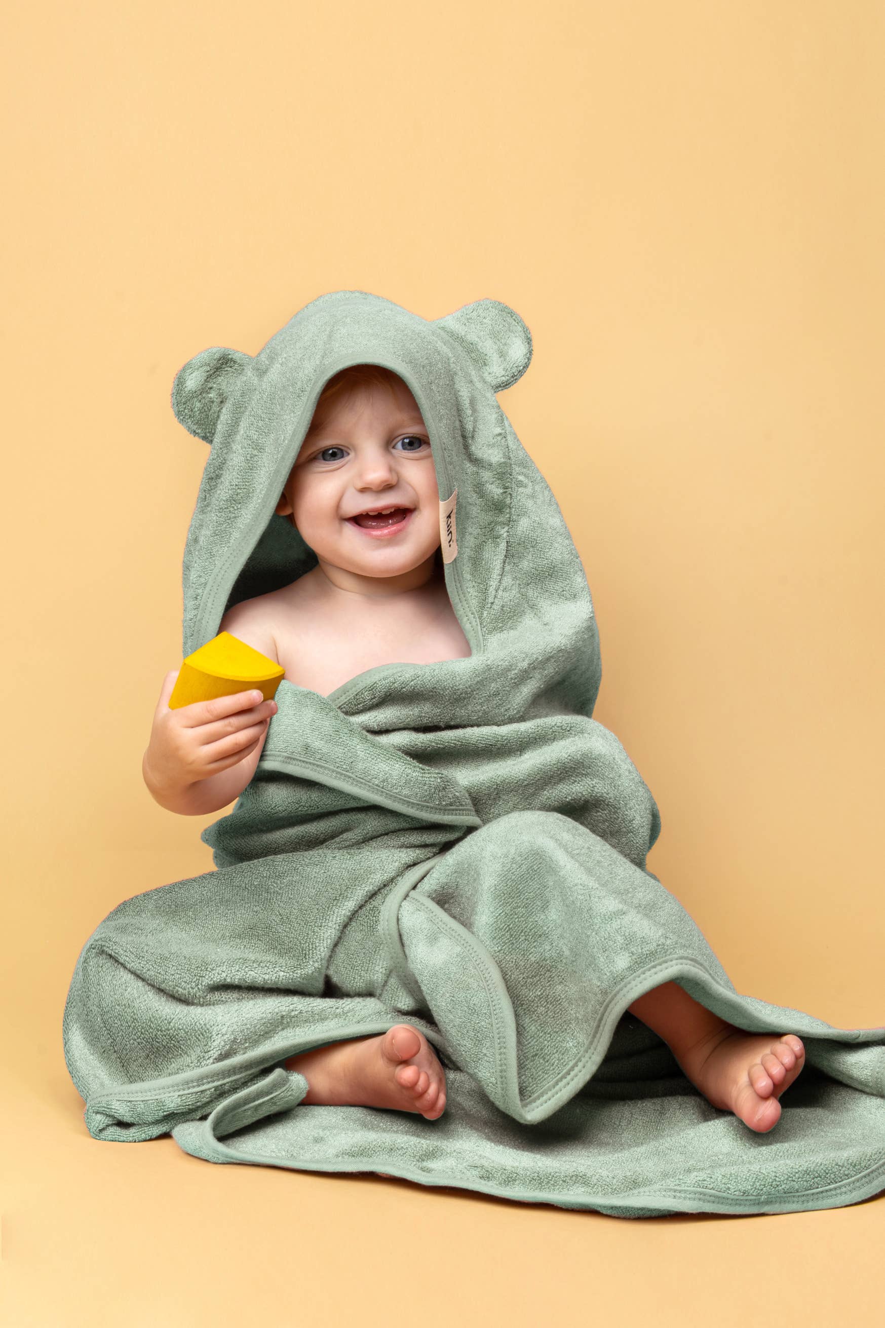 A baby smiles wrapped in a KIIN BABY Hooded Towel Ivory with bear ears, sitting on a yellow background while holding a yellow toy block.