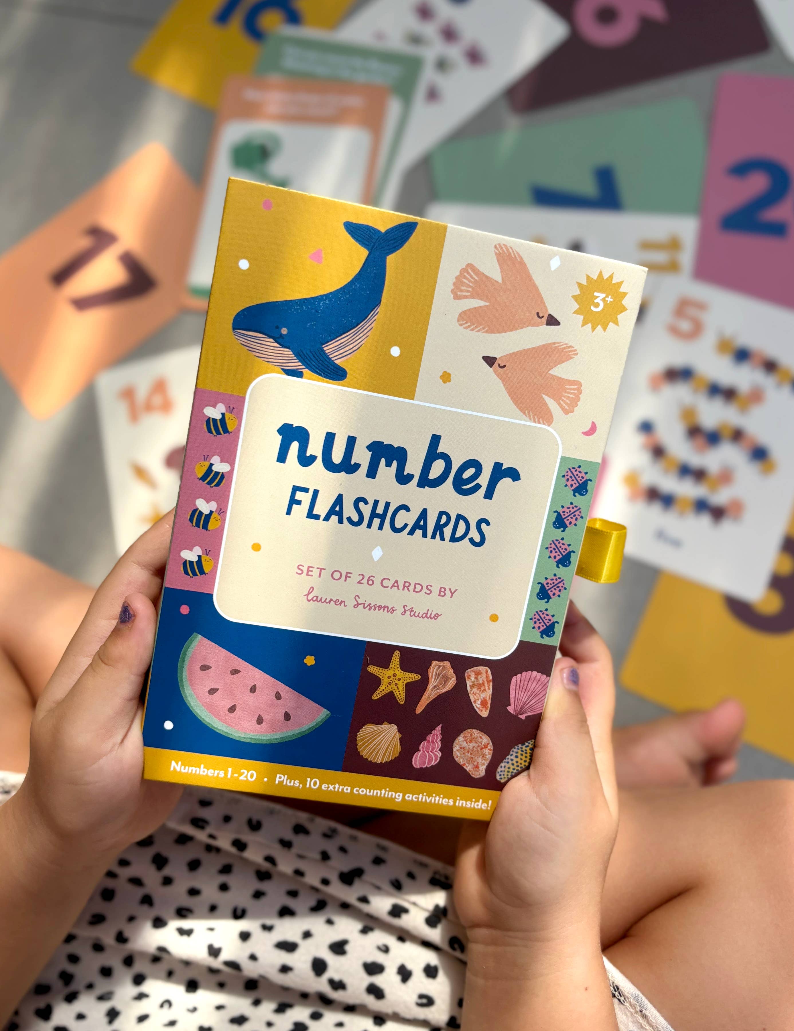 A child holds a box of Lauren Sissons - Illustrated Number Flashcards by Lauren Sissons Studio, designed to build counting skills with vibrant images of whales, birds, and objects. Some learning cards are spread out on the surface behind.