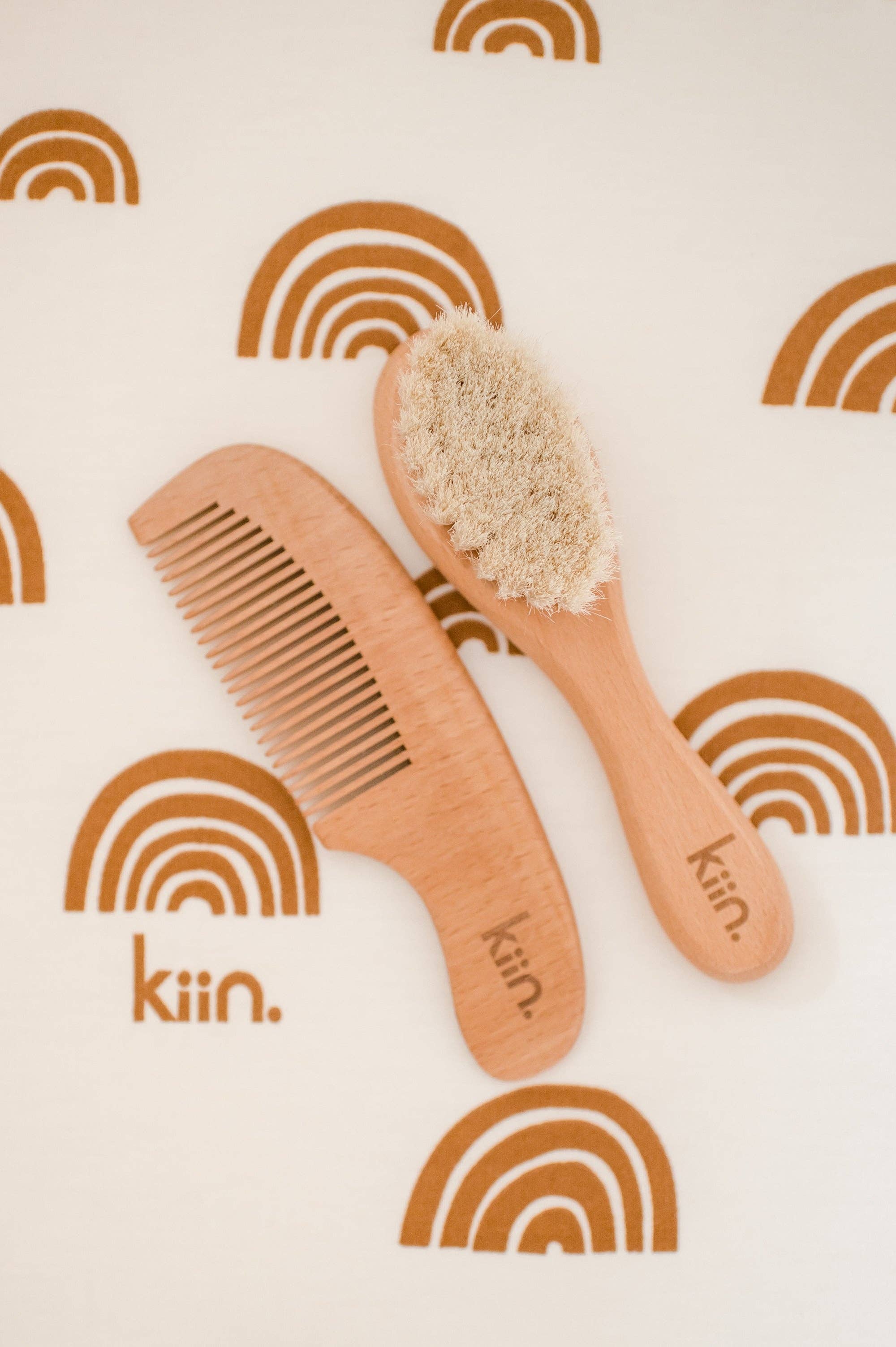 Wooden Baby Brush and Comb Set by KIIN BABY on white fabric with brown rainbow patterns, crafted from eco-friendly goat wool. The brush features soft bristles for gentle care, engraved with "KIIN.