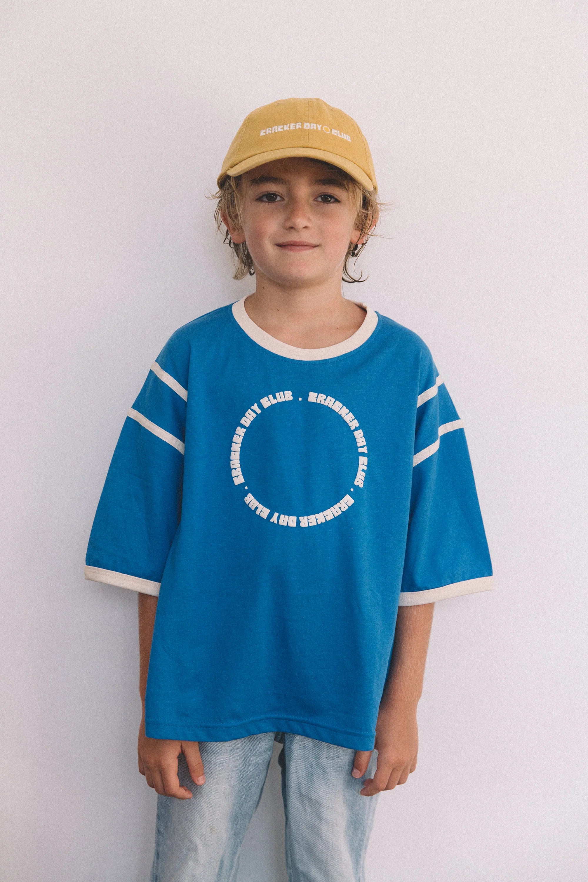 A child stands against a plain white wall, wearing the Cracker Day Club - Kids Racer Club T-Shirt Blue by CRACKER DAY CLUB, paired with light blue jeans and a yellow baseball cap—a playful update to classic outdoor style.