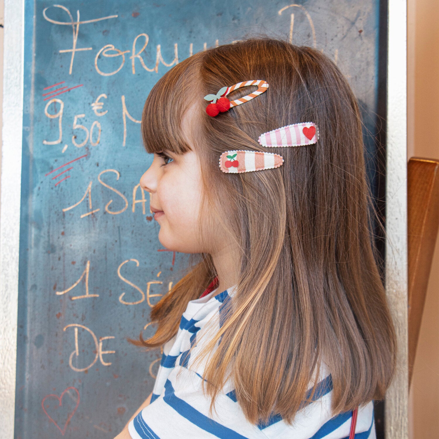 A young girl with brown hair wearing a striped shirt and ROCKAHULA's Rockahula - Sweet Cherry Clips stands in profile in front of a chalkboard with French writing and prices.