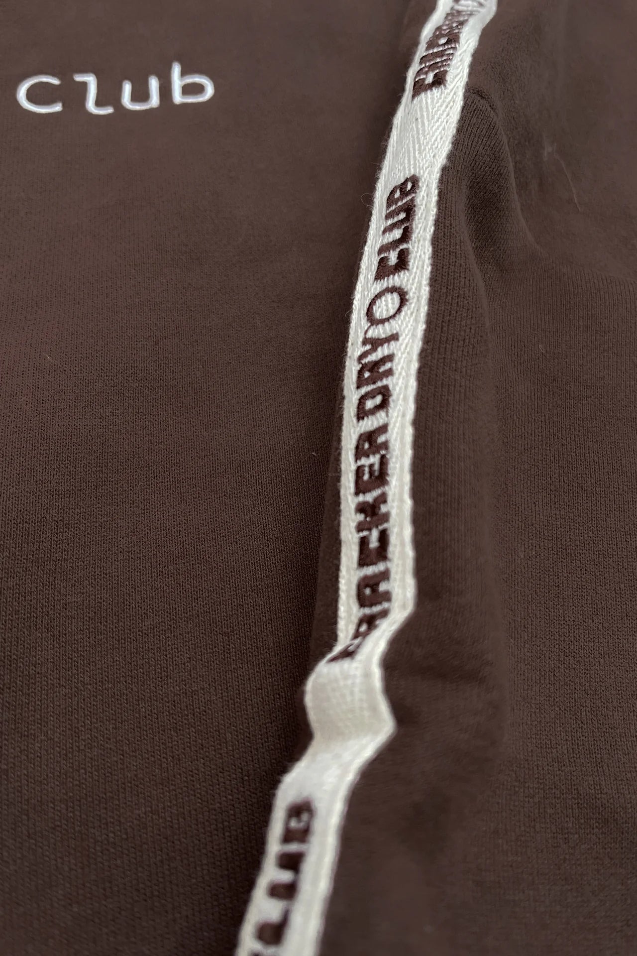 Close-up of heavyweight terry cotton in chocolate brown with “club” embroidered in white and a trim featuring “PLAYERVDNCLUB.” A perfect detail from CRACKER DAY CLUB’s Cloud Racer Hoodie Chocolate collection.