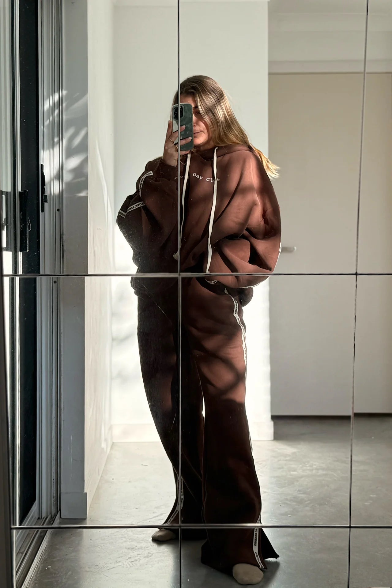 Wearing CRACKER DAY CLUB's Cloud Racer Sweatpants in Chocolate, a person snaps an indoor mirror selfie, phone obscuring their face. Sunlight pours in from the left, casting shadows across the wall and heavyweight terry cotton floor.