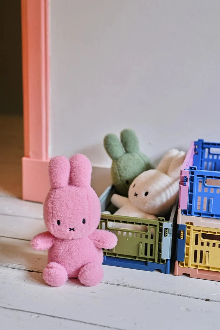 A Miffy ECO Cotton Candy Rose 23cm by BON TON TOYS X MIFFY rests on a white wooden floor near colorful plastic crates. Inside the crates are partially visible plush toys, one green and one white. The simple setting features a light-colored wall and a pink doorway.