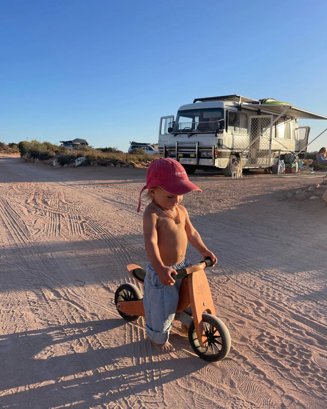 A young child wearing a red cap and ZIGGY ZAZA’s Suki Denim Jeans in Light Wash rides a wooden balance bike on a dirt road, with an RV and campsite in the background under a clear blue sky.