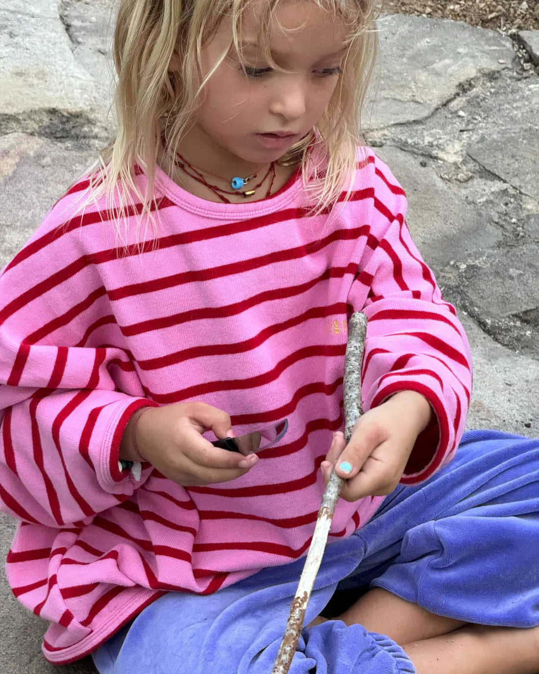 A young child in a ZIGGY ZAZA Row Row Pullover Rose Stripe sits outside on a stone surface, carefully whittling a stick with a small knife.