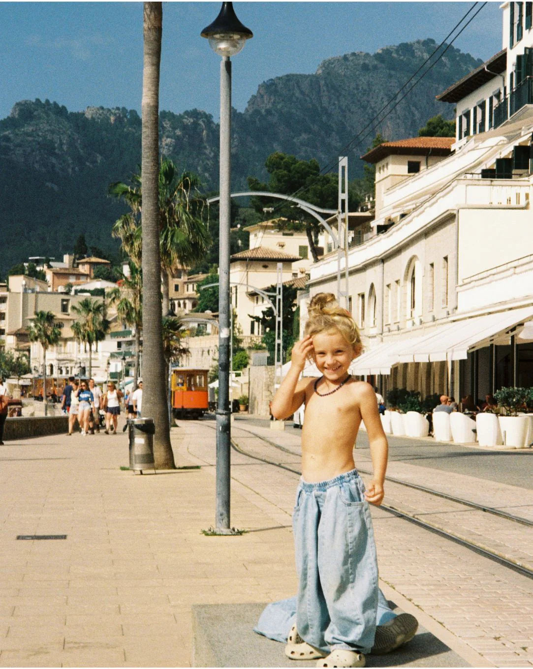 A young child smiles on a sunny beach promenade wearing ZIGGY ZAZA’s Ziggy Zaza - Suki Denim Jean Light Wash Denim and Crocs, with palm trees, mountains, and Mediterranean-style buildings in the background.