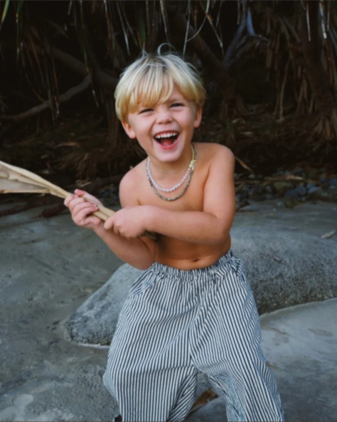 A young blonde boy stands outdoors, laughing joyfully while wearing the Ziggy Zaza - Casper Pant Navy Stripe by ZIGGY ZAZA. He is shirtless and holding a wooden paddle, surrounded by rocks and dense greenery.