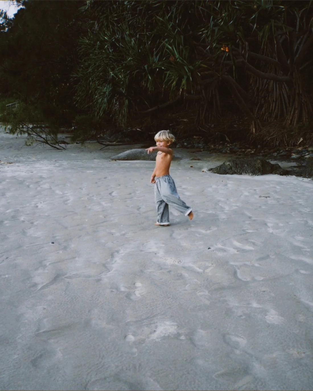 A young, shirtless child with light hair walks barefoot on a sandy beach, wearing relaxed fit Ziggy Zaza - Casper Pant Navy Stripe by ZIGGY ZAZA, pointing into the distance with dense greenery in the background.