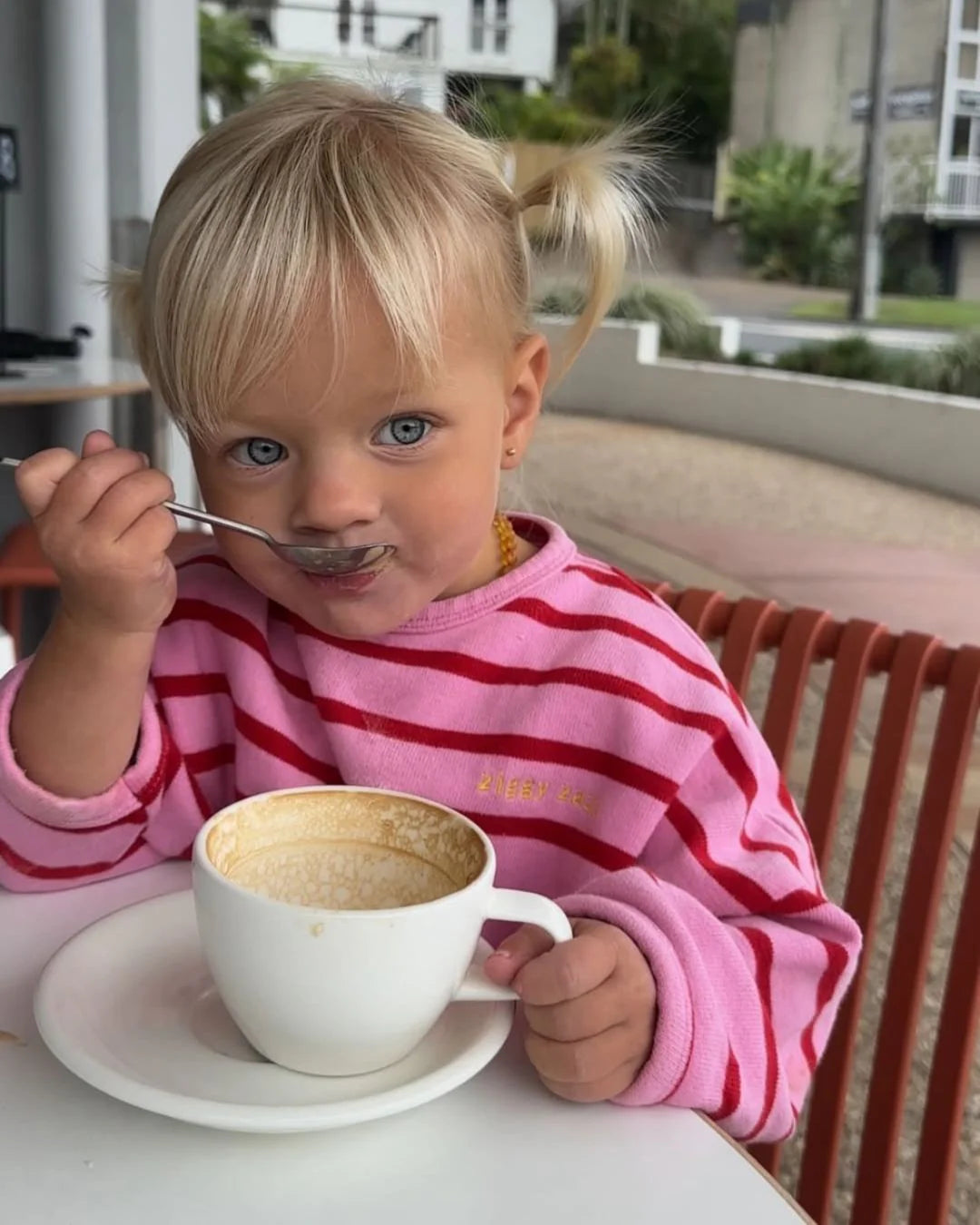 A young blonde-haired child in pigtails wears the Ziggy Zaza - Row Row Pullover Rose Stripe by ZIGGY ZAZA, holding a large white cup with froth and eating with a spoon at an outdoor table.