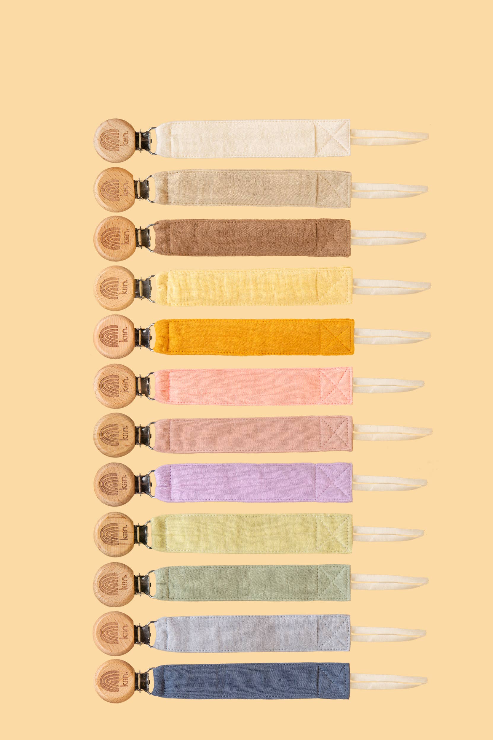 A vertical row of twelve KIIN BABY Cotton Dummy Clips features various colors transitioning from neutral tones at the top to bright pastels in the middle and darker hues at the bottom. Each has a round wooden fastener, ideal for any nursery or baby shower gift, set against a soft yellow background.