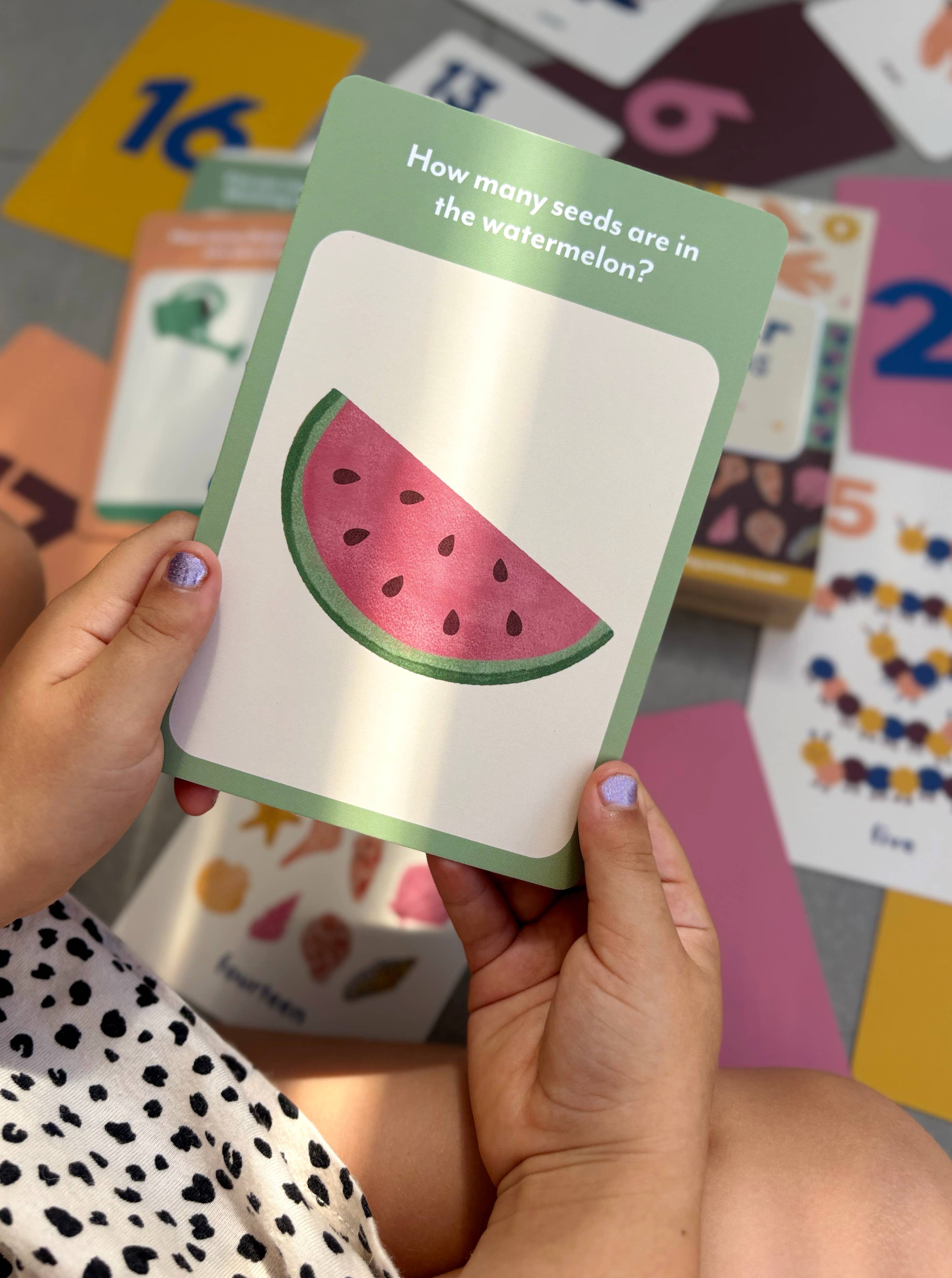 A child holds a Lauren Sissons - Illustrated Number Flashcard with a watermelon slice and the question, "How many seeds are in the watermelon?" Colorful Lauren Sissons Studio cards are scattered behind, encouraging counting skills.