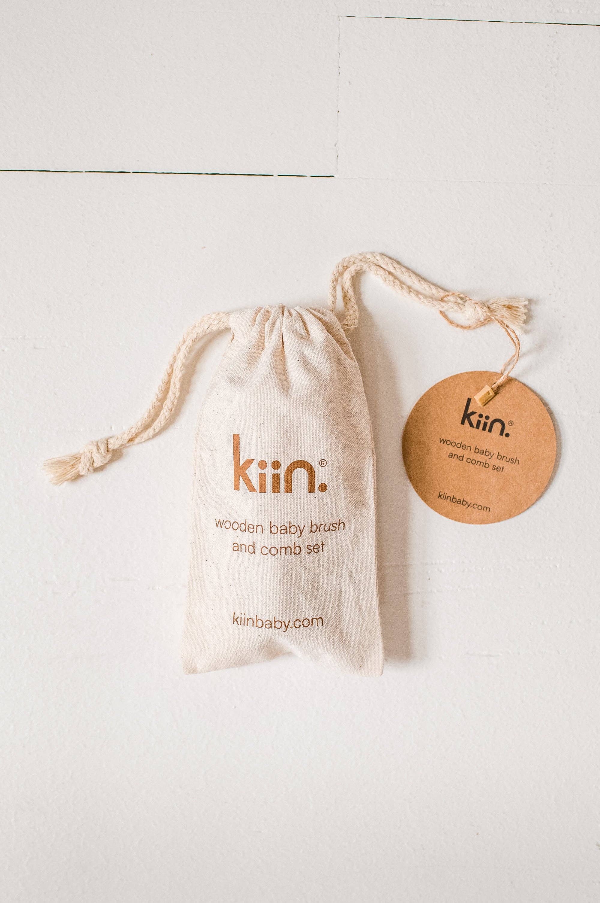 A beige drawstring bag with "KIIN BABY" and "Wooden Baby Brush and Comb Set" on it, made from eco-friendly materials, is adorned with a circular tag against a white background. The set includes a baby hair brush crafted from soft goat wool for gentle care.