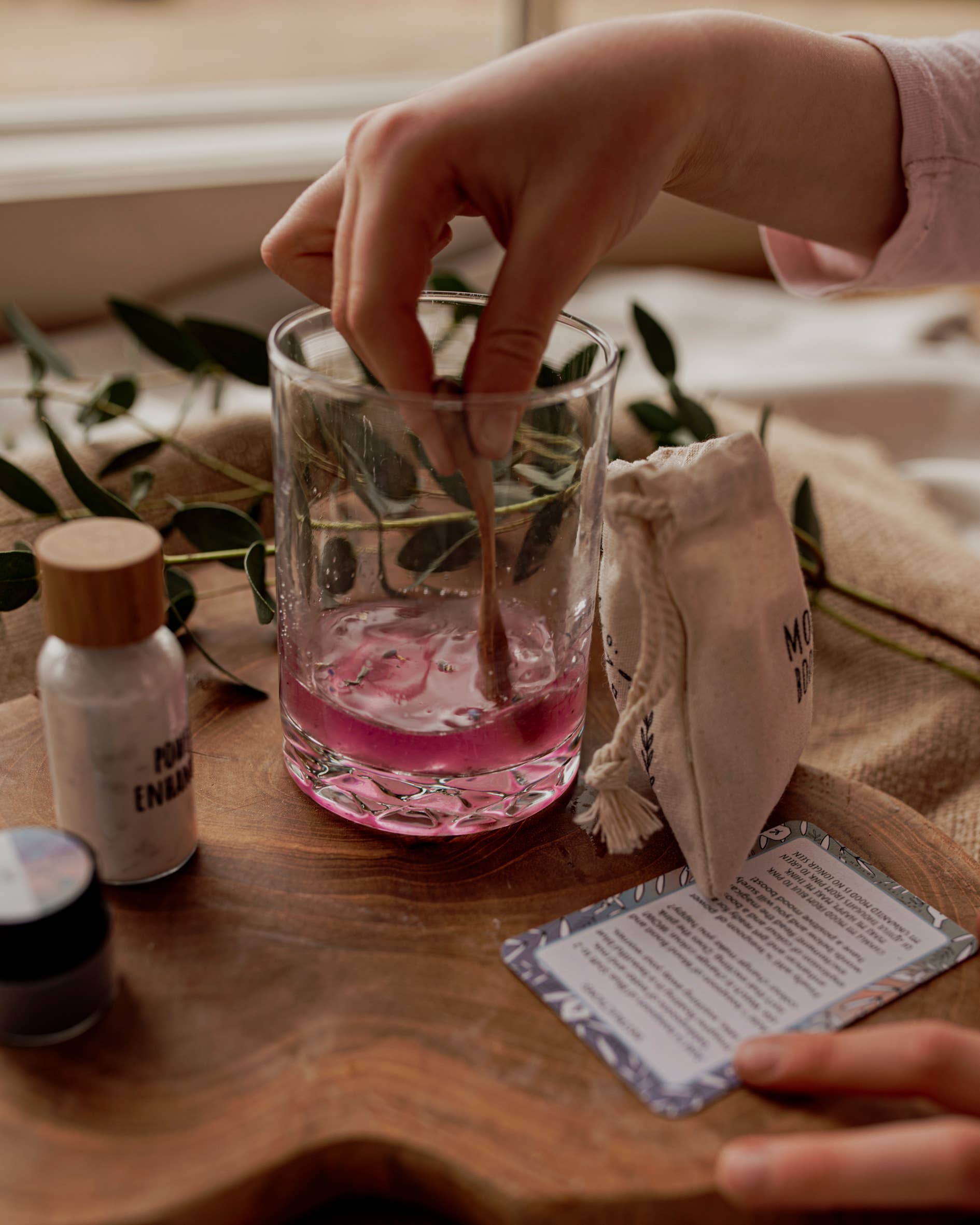 A hand stirs pink liquid in a glass using a wooden spoon, reminiscent of The Little Potion Co's MINI Colour Mood Potion Kit. Nearby are small containers, a cloth bag, and a card. Eucalyptus leaves add to the enchanting sensory play experience by THE LITTLE POTION CO.