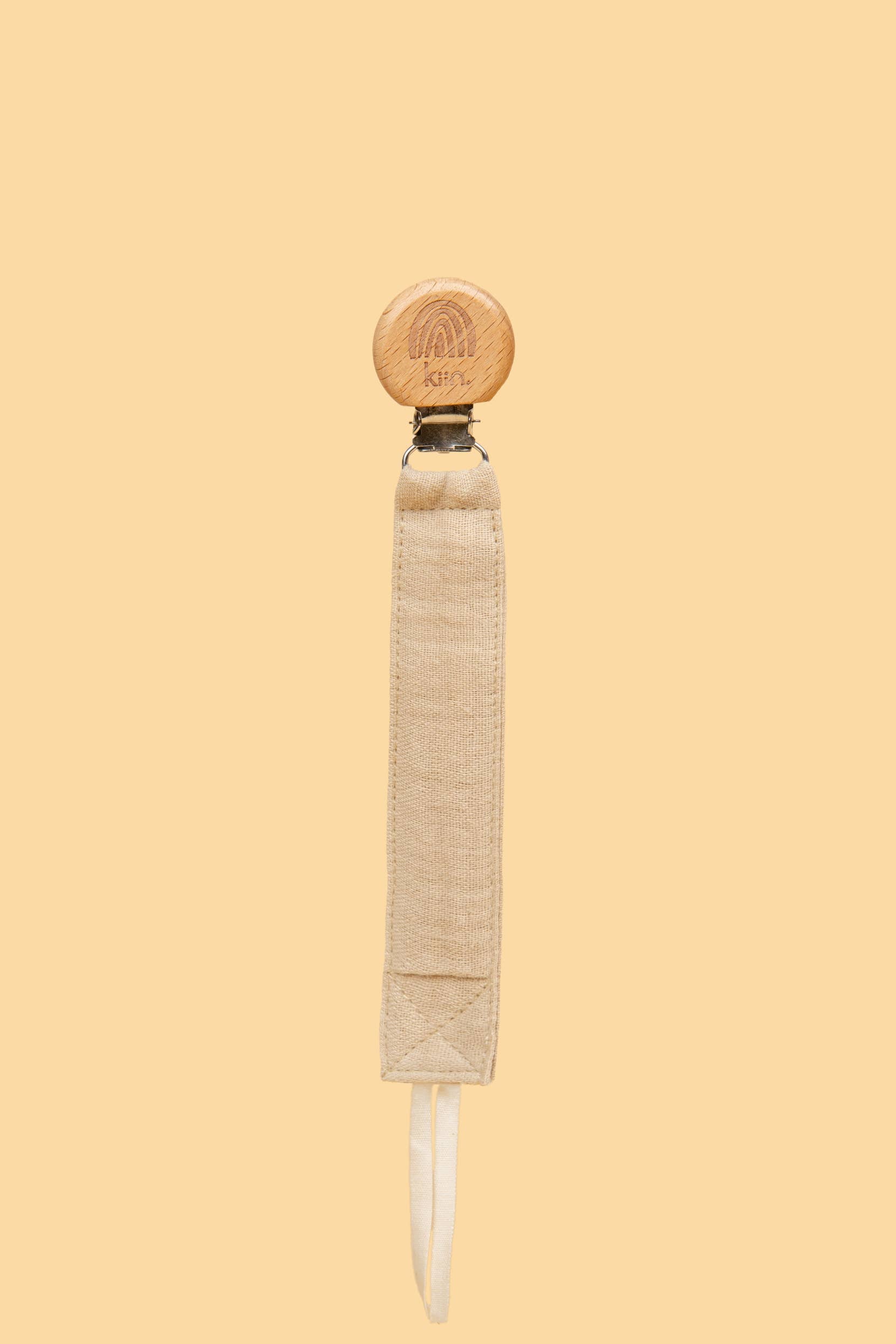 The KIIN BABY Cotton Dummy Clip Oat features a wooden clip with a small rainbow engraving and a beige cotton strap displayed on a light yellow background, making it perfect for gender-neutral nurseries or as an ideal baby shower gift.