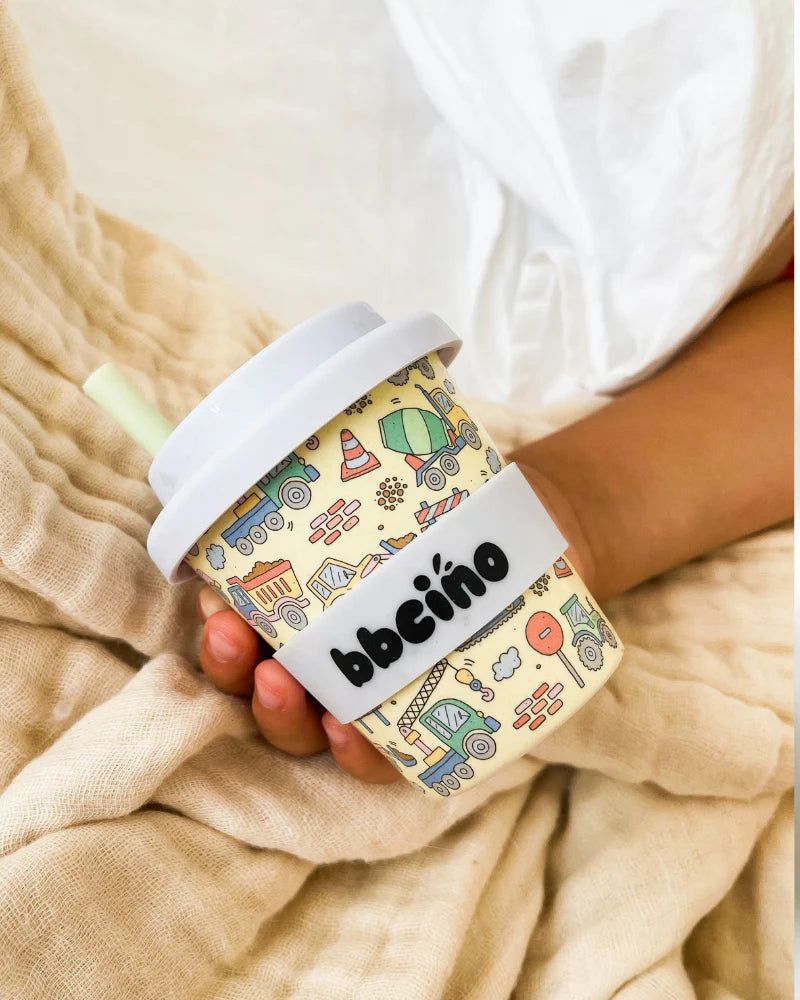 A hand holds the BBCINO BBcino ~ Reusable Babycino Cup 120ml Dig It, an eco-friendly bamboo cup with a white lid and straw. The cup features illustrations of trucks and traffic cones with "bbčīno" on a white band, set against beige fabric.