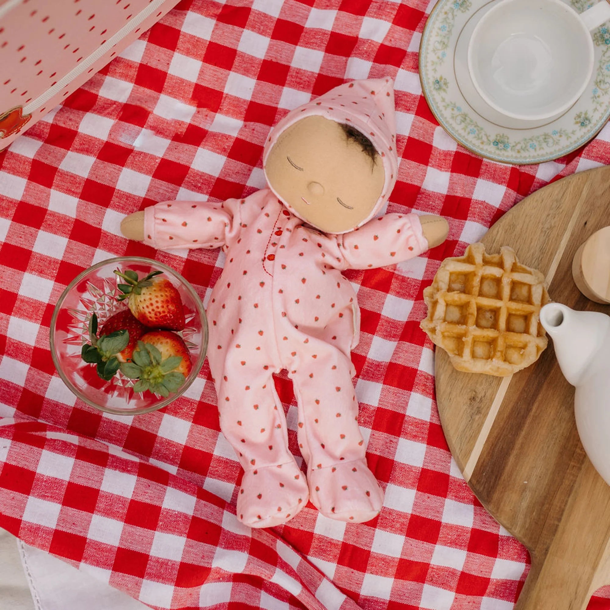 A child's Dozy Dinkum Strawberry Pip doll from OLLI ELLA, dressed in pink polka dots, lies on a red and white checkered picnic blanket. Surrounding the OLLI ELLA doll are a bowl of strawberries, a waffle, and tea set pieces on a wooden tray. The scene conveys a playful picnic setting.