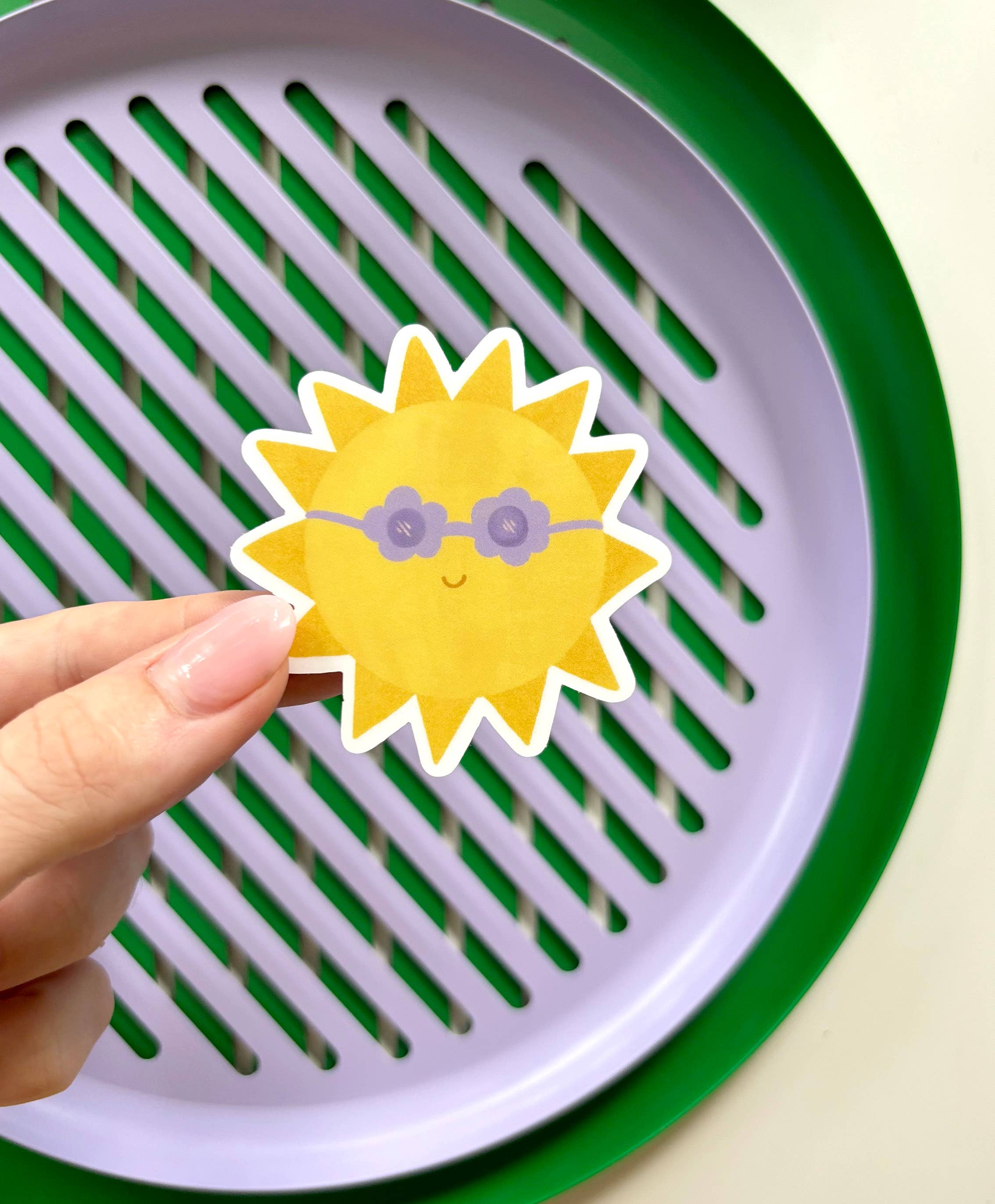 A hand holding the cheerful Sunshine Sticker by LAUREN SISSONS STUDIO, featuring a cartoonish sun with purple sunglasses. The background showcases a round, lavender-colored tray with green trim and radial slotted vents, spreading positive vibes. This weatherproof vinyl sticker brightens any day!