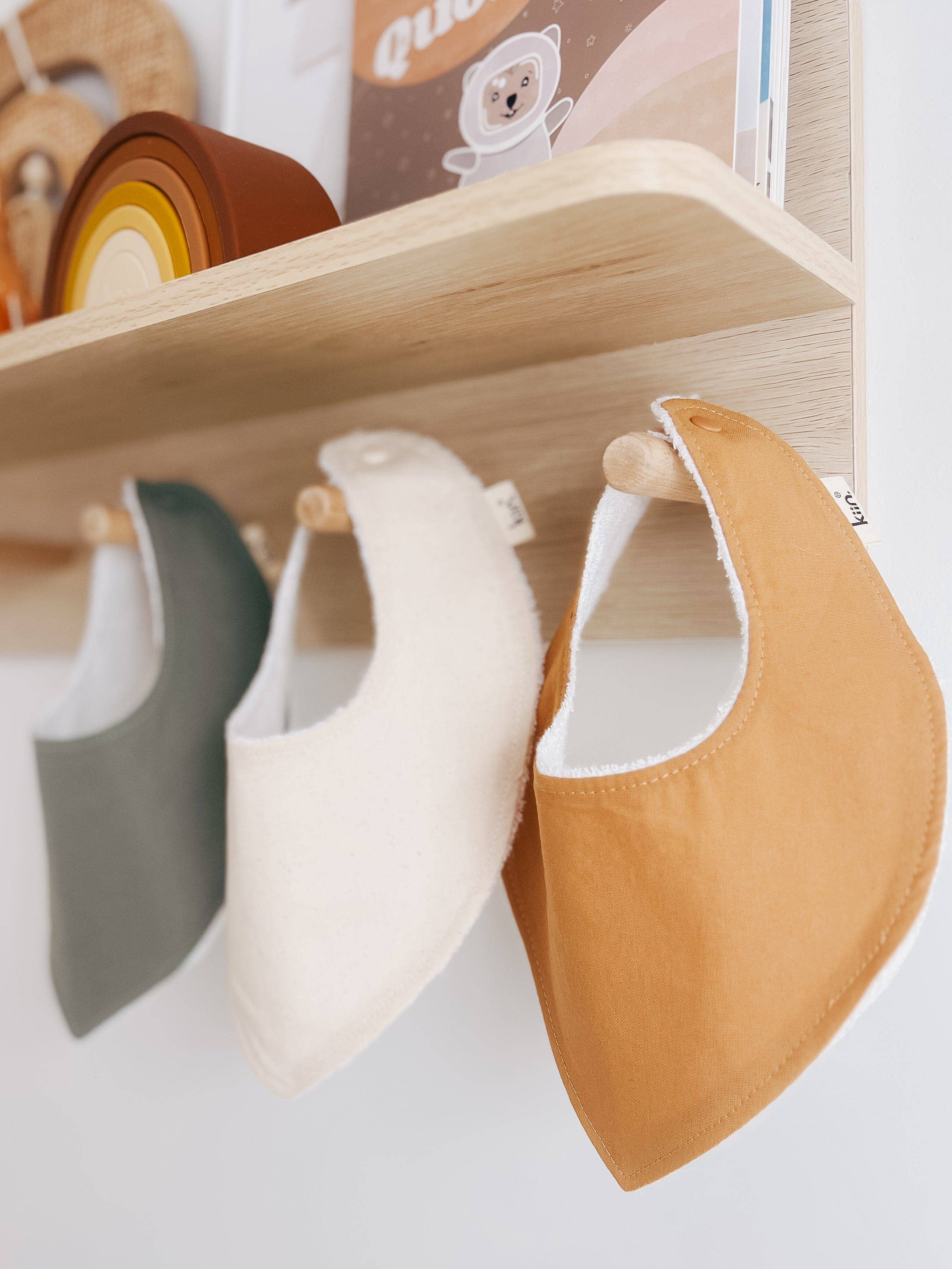 Three KIIN BABY Dribble Bibs in green, cream, and orange, crafted from soft bamboo/cotton towelling with snap fasteners, hang from wooden hooks beneath a shelf. The shelf above holds children's books and toys, including a rainbow stacking toy. The setup is against a white wall.