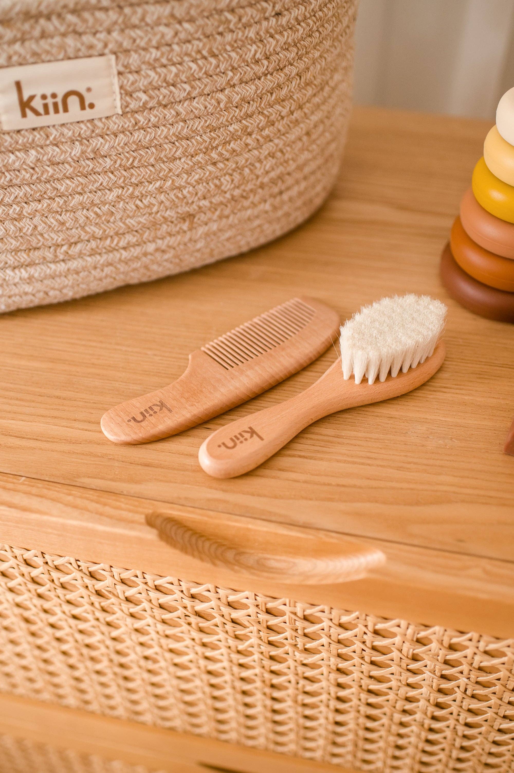 On a wicker basket surface, the Wooden Baby Brush and Comb Set by KIIN BABY features eco-friendly, softly lit goat wool bristles. A stack of colorful rings and a woven basket enhance the cozy scene.