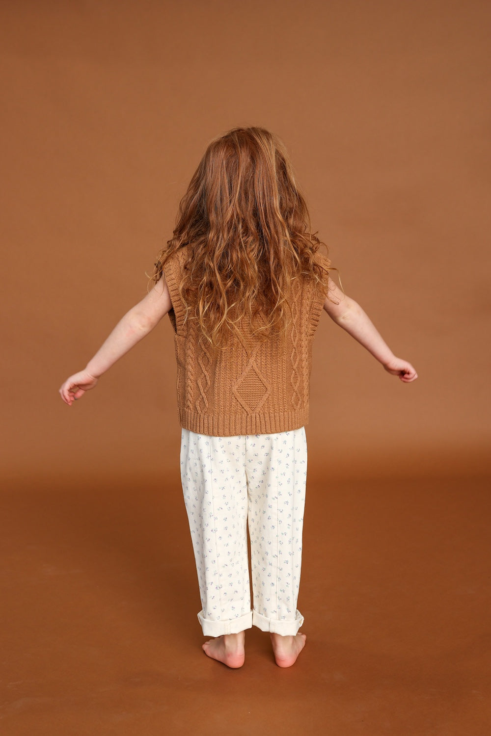 A young child with long, wavy brown hair stands facing away, wearing GROWN's Everyday Pants Garden—relaxed fit with a small print and cotton elastic waistband—paired with a brown knit vest, set against a plain brown background.