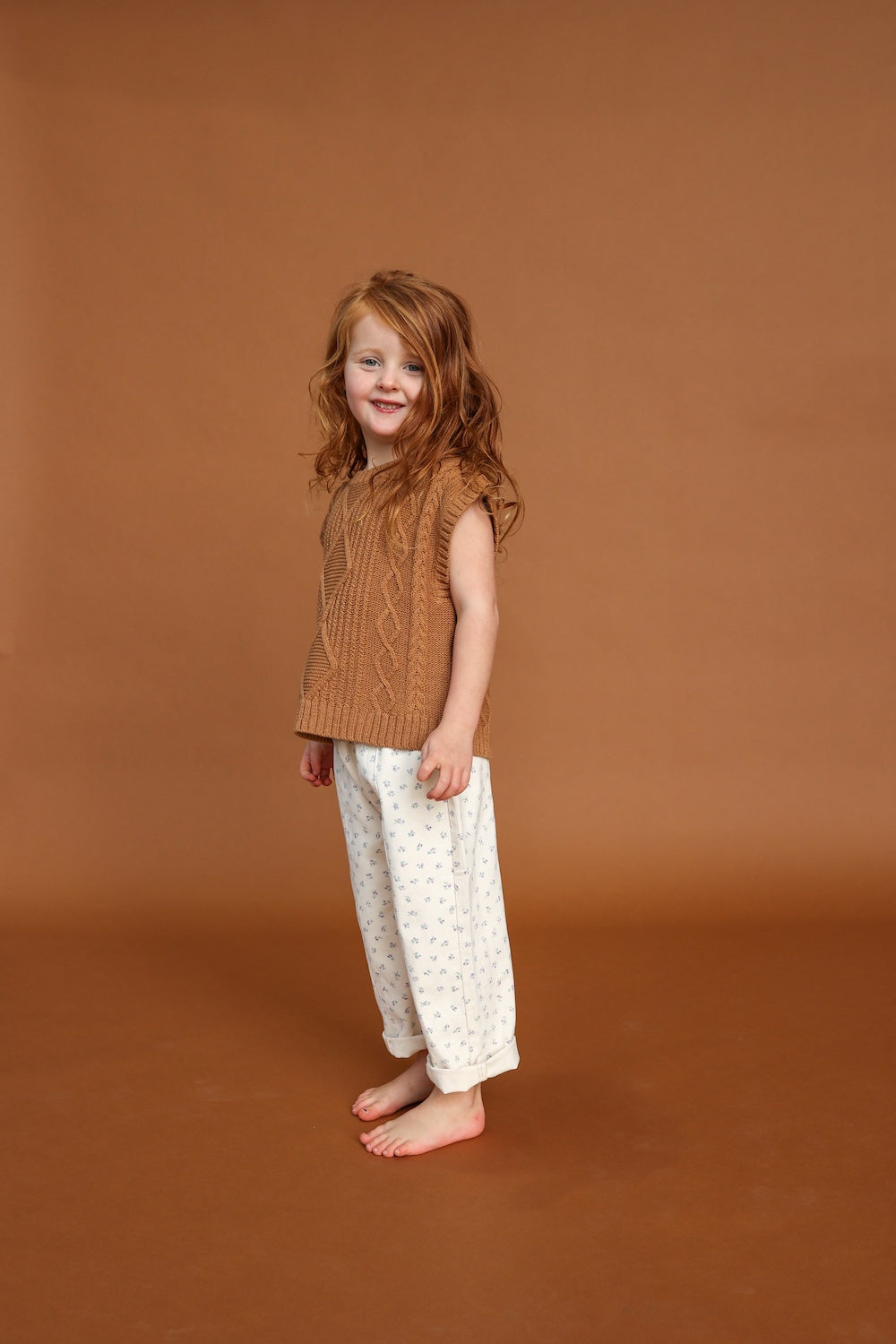 A young, red-haired child smiles barefoot on a brown floor and background, wearing a brown knitted sleeveless sweater and GROWN’s Everyday Pants Garden by GROWN, featuring a subtle blue pattern.