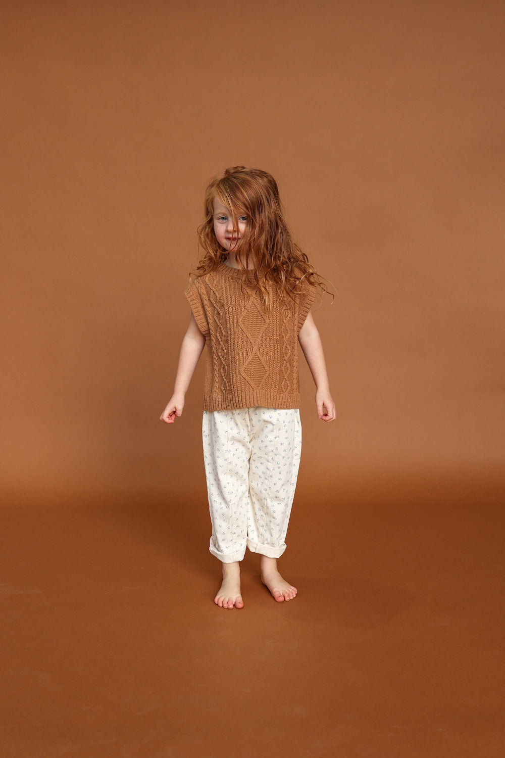 A young child with long red hair stands barefoot on a brown floor, wearing GROWN's Everyday Pants Garden—relaxed fit pants with a subtle pattern and cotton elastic waistband—in front of a plain brown background.